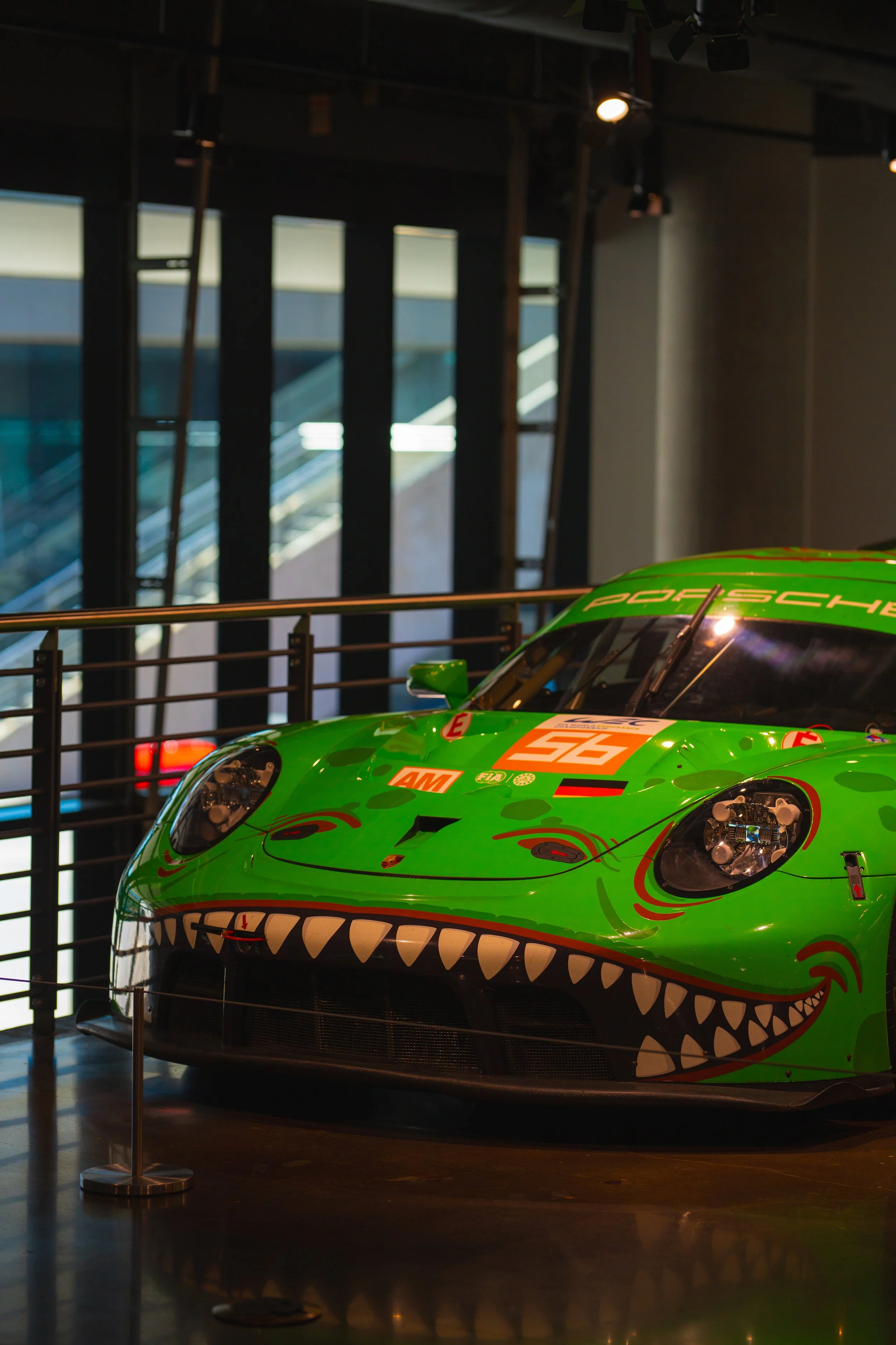 Green Porsche race car with a cartoonish face and shark teeth design, displayed indoors behind a black railing.