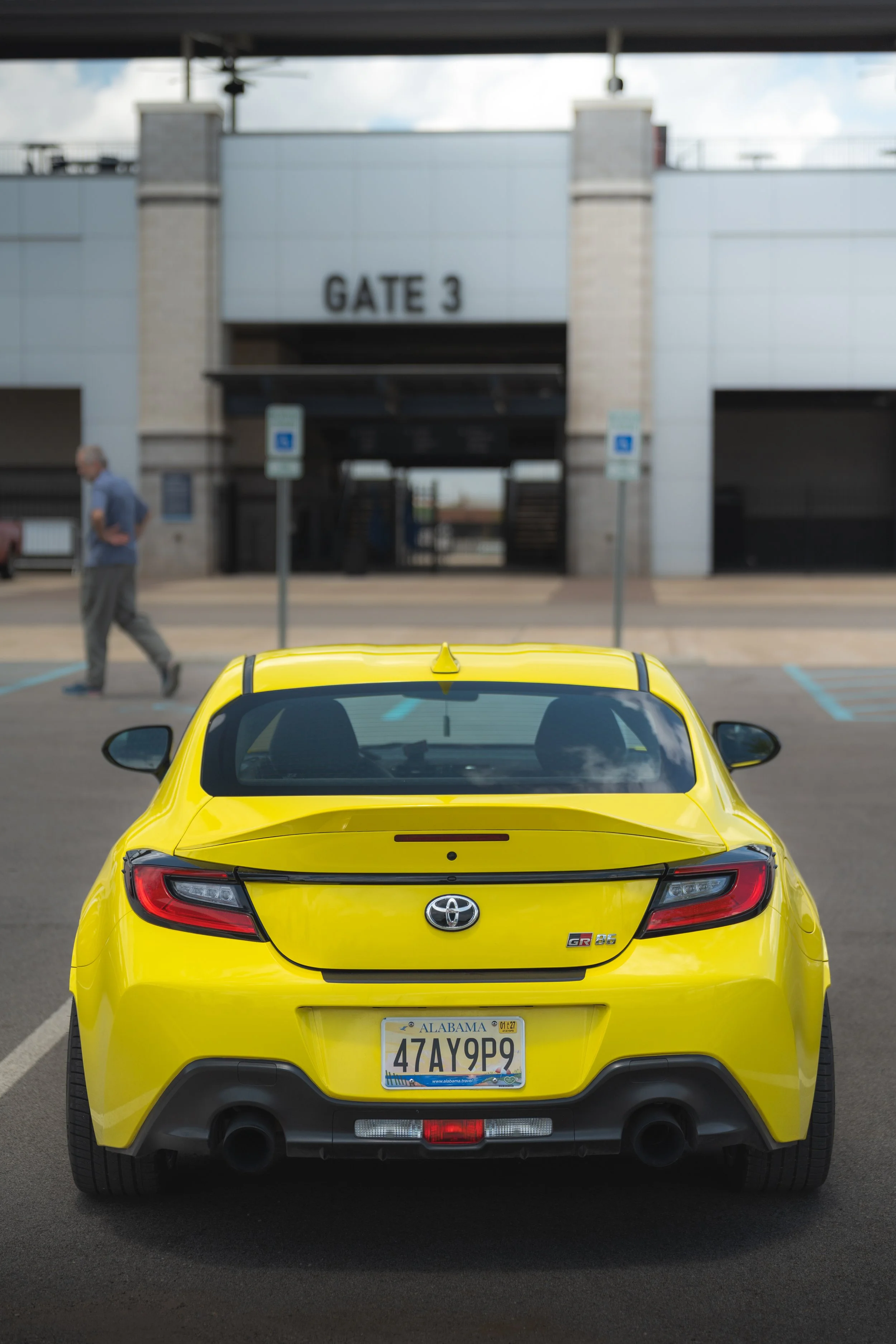 rear view of a yellow Toyota GR 86 sports car with Alabama license plate 47AY9P, parked in a parking lot facing a building with a gated entrance labeled 'GATE 3'.