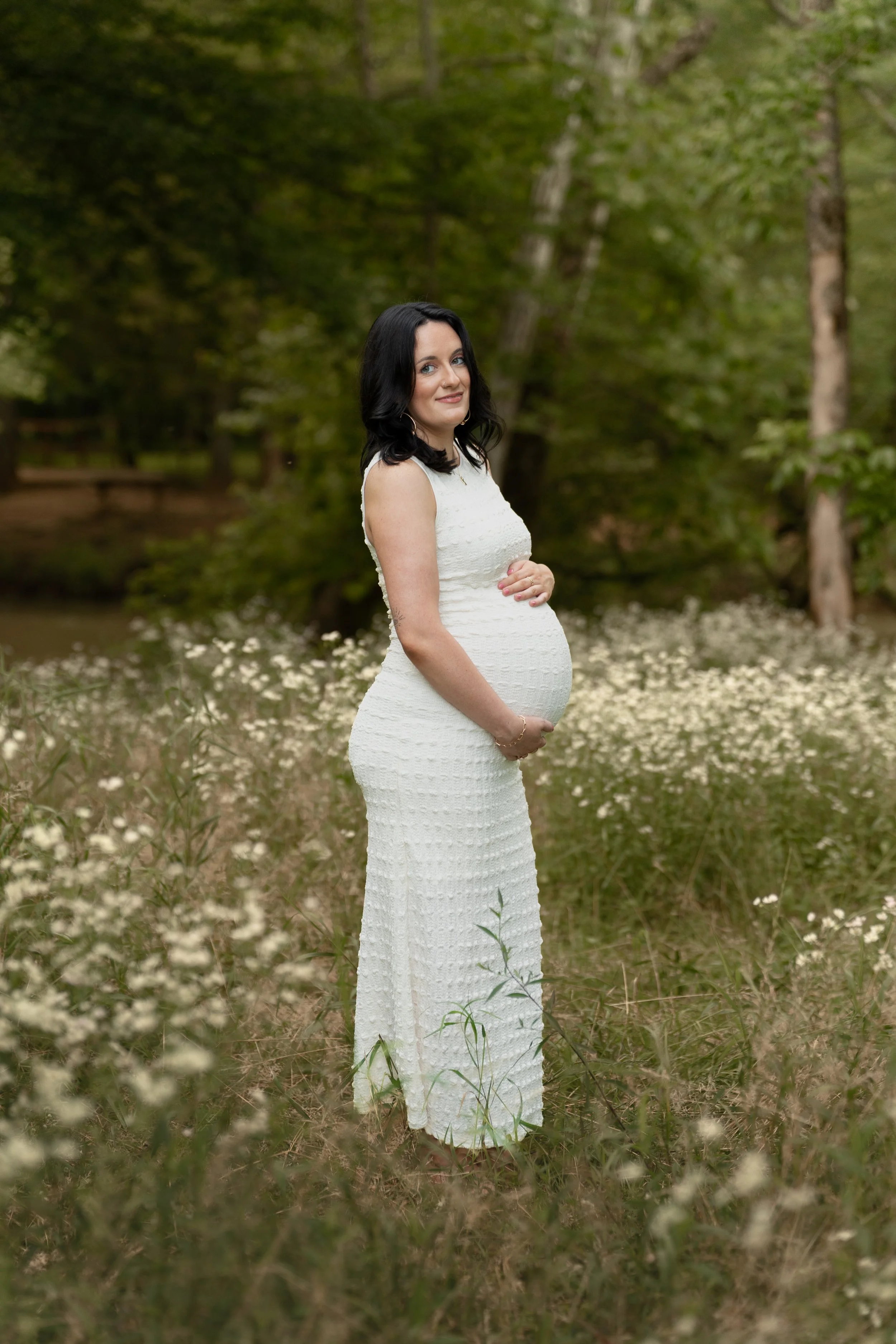 A pregnant woman with black hair, wearing a white dress, standing in a field of small white flowers near a pond, with trees in the background.