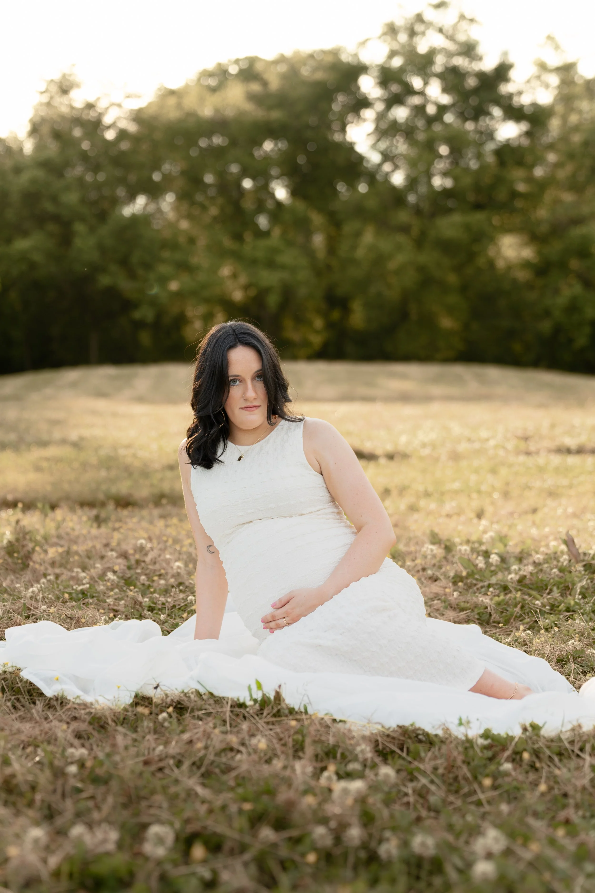 A woman with dark hair sitting on the grass in a white dress, outdoors during sunset with trees in the background.