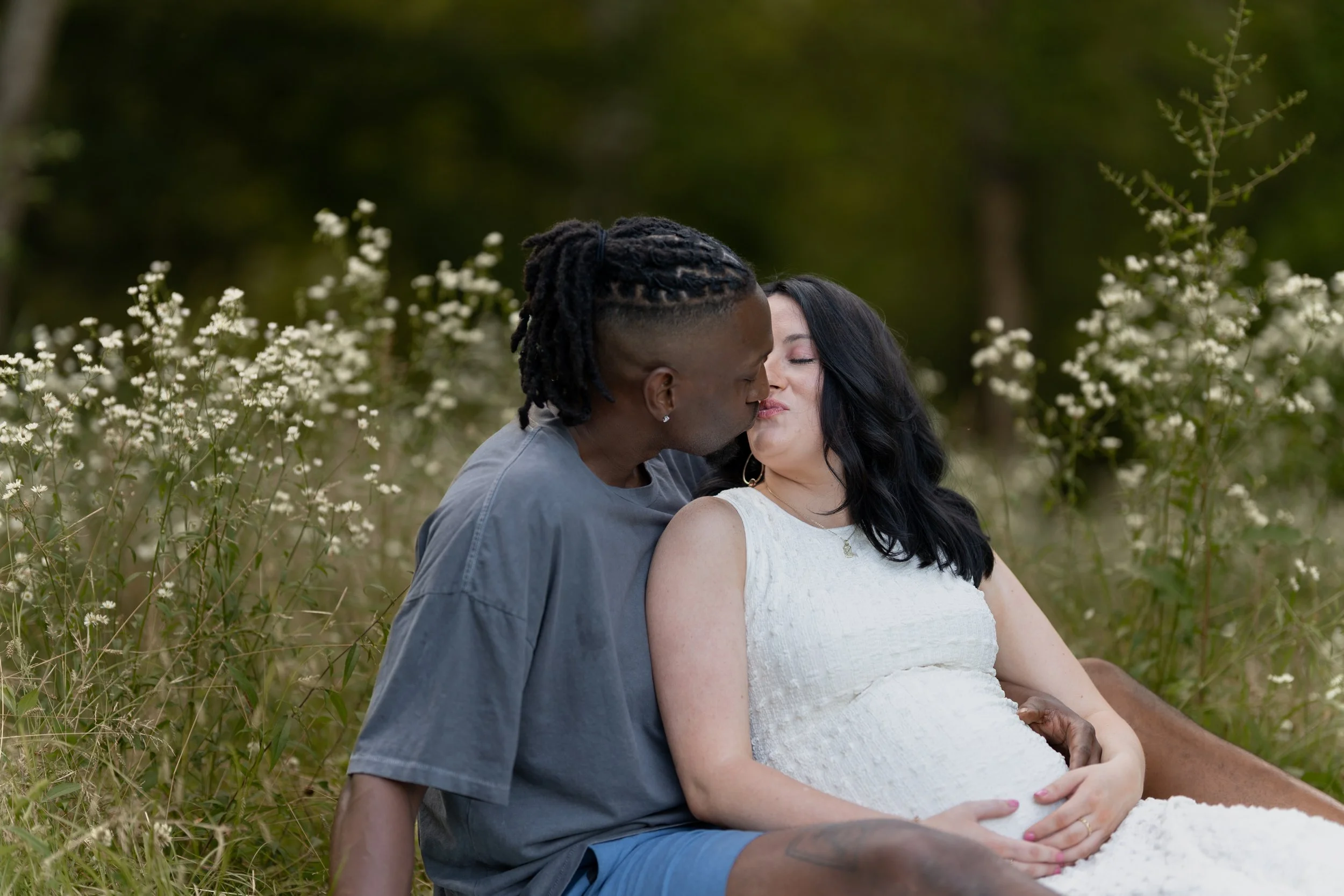 A couple sharing a kiss in a grassy outdoor setting with white flowers and green trees.