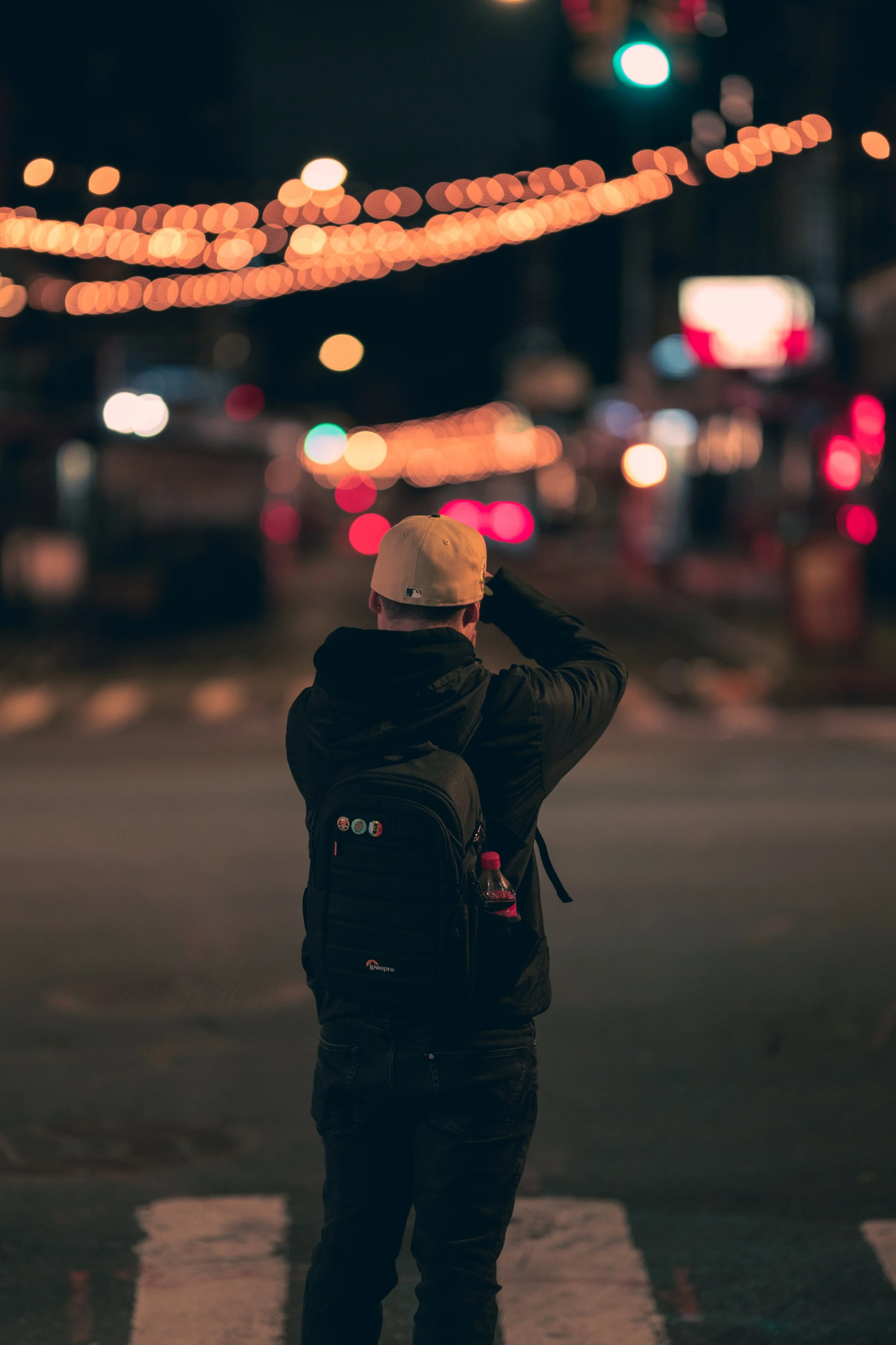 Person standing on city street at night, seen from behind, wearing a beige cap, black jacket, backpack with pins, and holding a water bottle, with blurred city lights and traffic in the background.