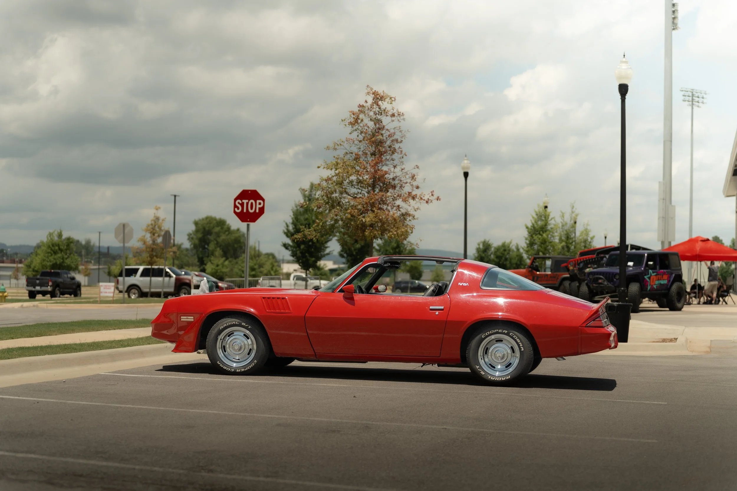 A red sports car parked in an outdoor lot on a cloudy day, with other vehicles, trees, a stop sign, and street lamps visible in the background.