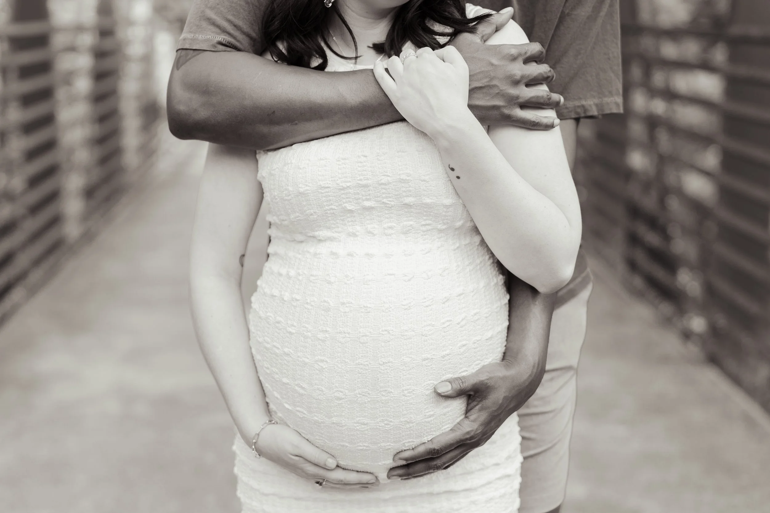 A pregnant woman in a white textured dress being embraced from behind by a man, with other arms wrapping around her, in an outdoor setting.