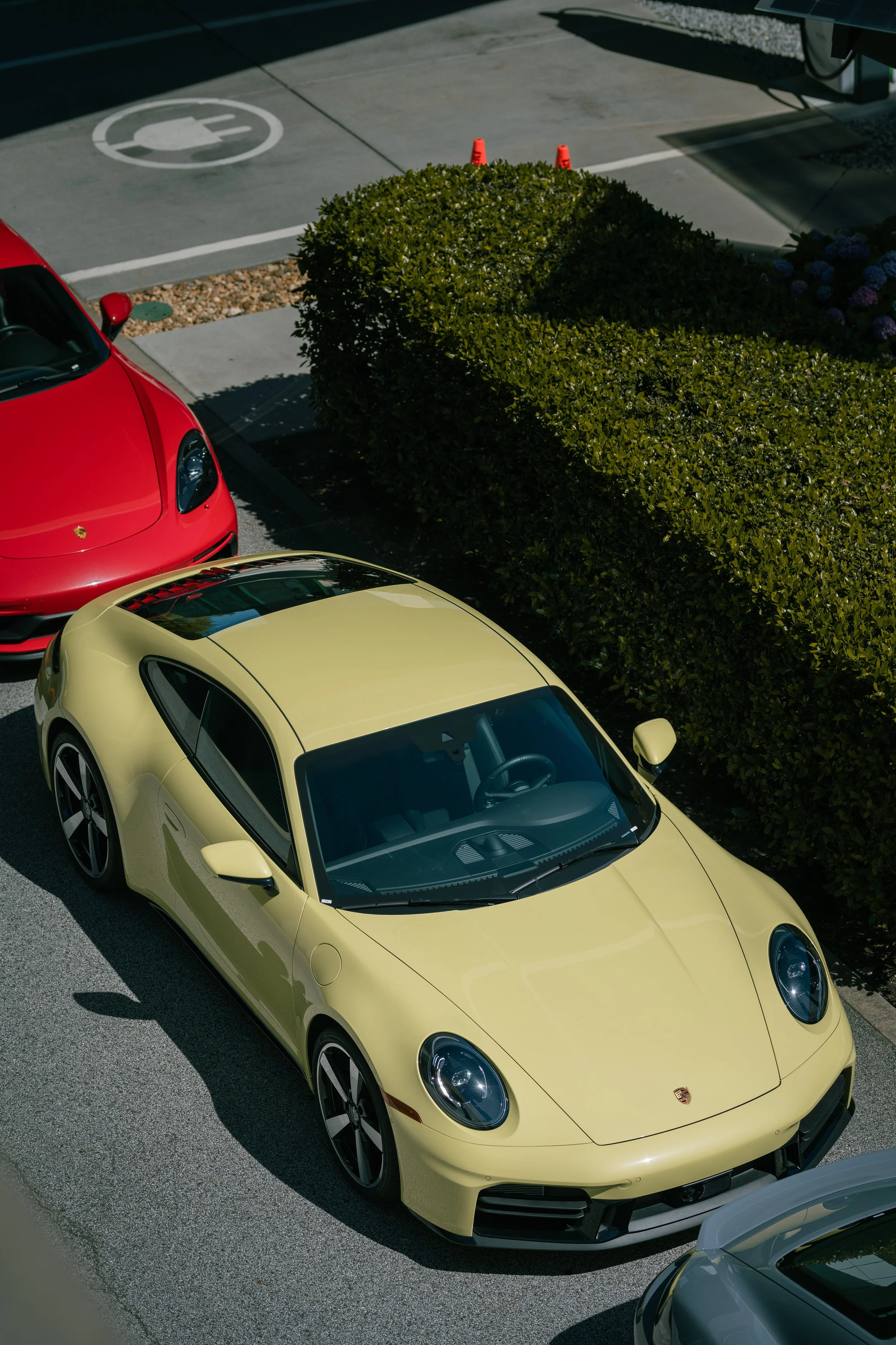 Yellow Porsche sports car parked next to a green hedge, with a red sports car partially visible behind it, and a parking space marked for electric vehicle charging in the background.