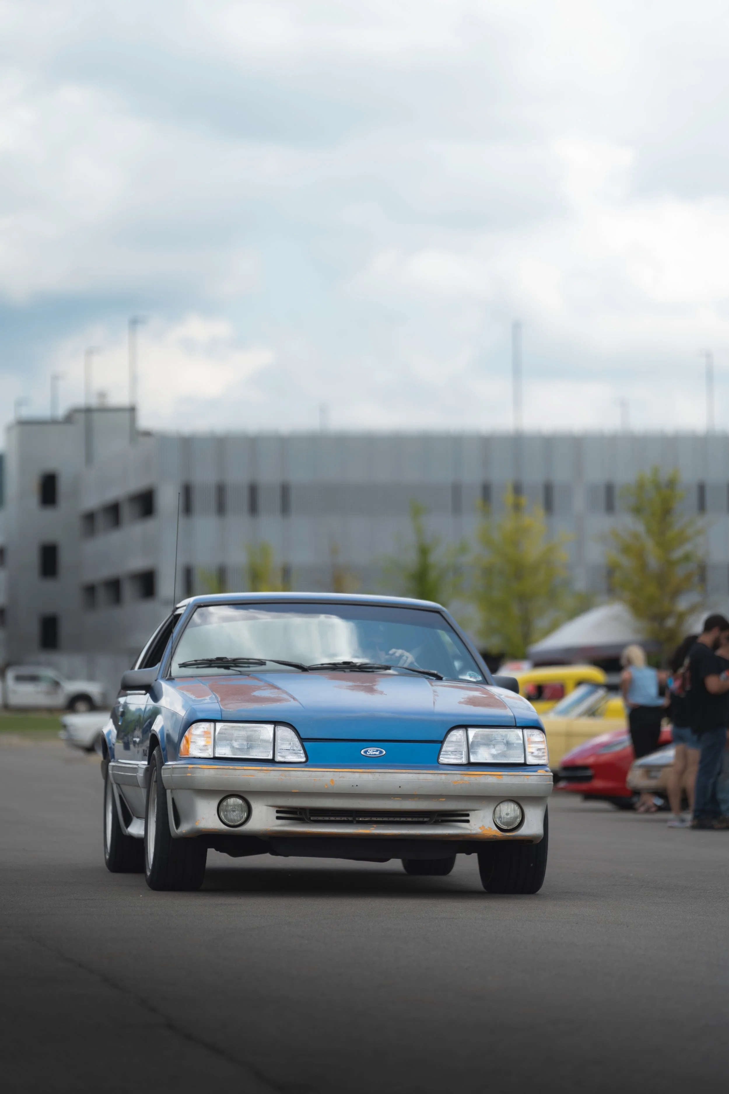 Blue and gray Ford Mustang car parked in the foreground at a car show, with people and other cars in the background, overcast sky, and a building behind.