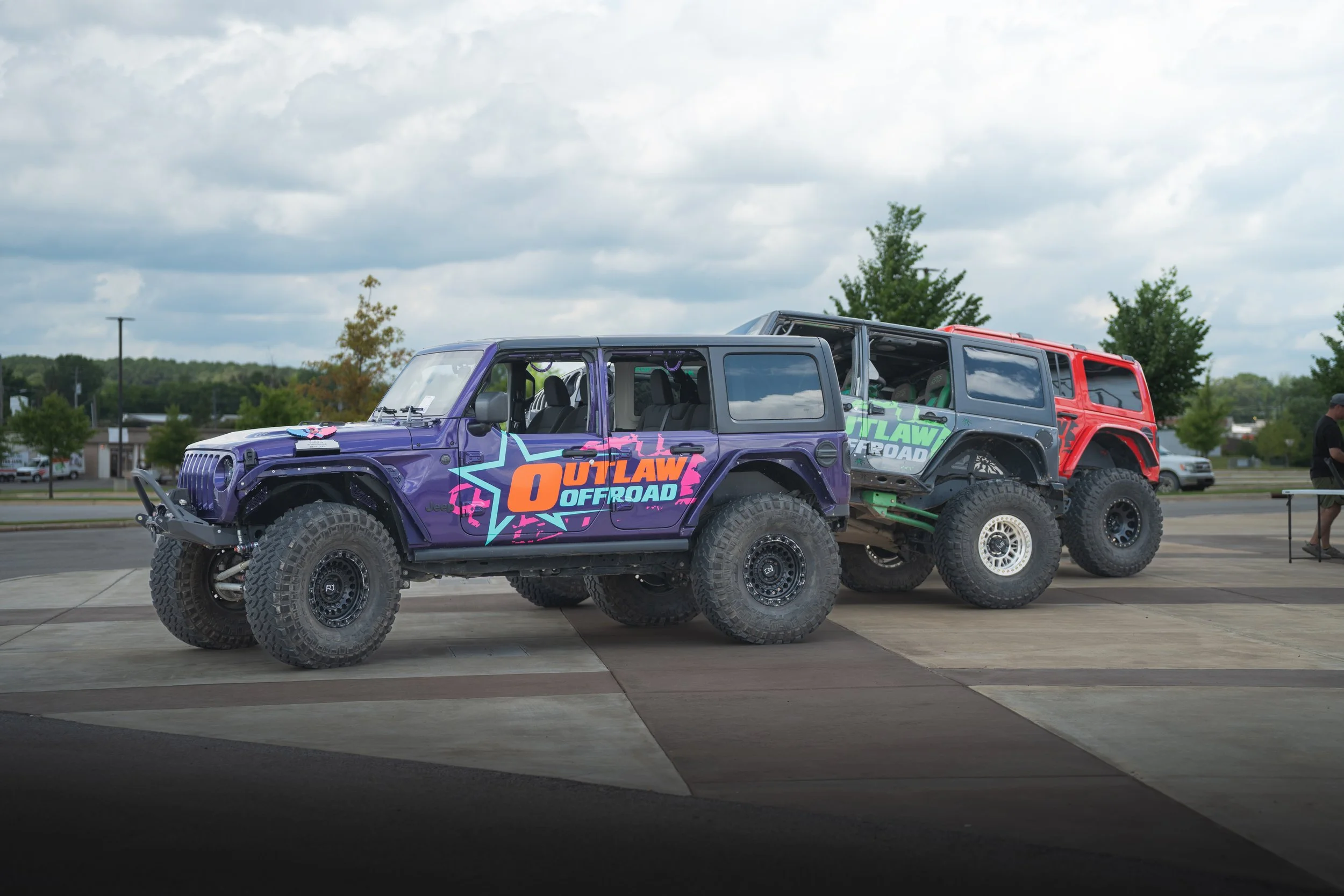 Two off-road vehicles parked outdoors, one purple and one red, with colorful branding and large tires, under a cloudy sky.
