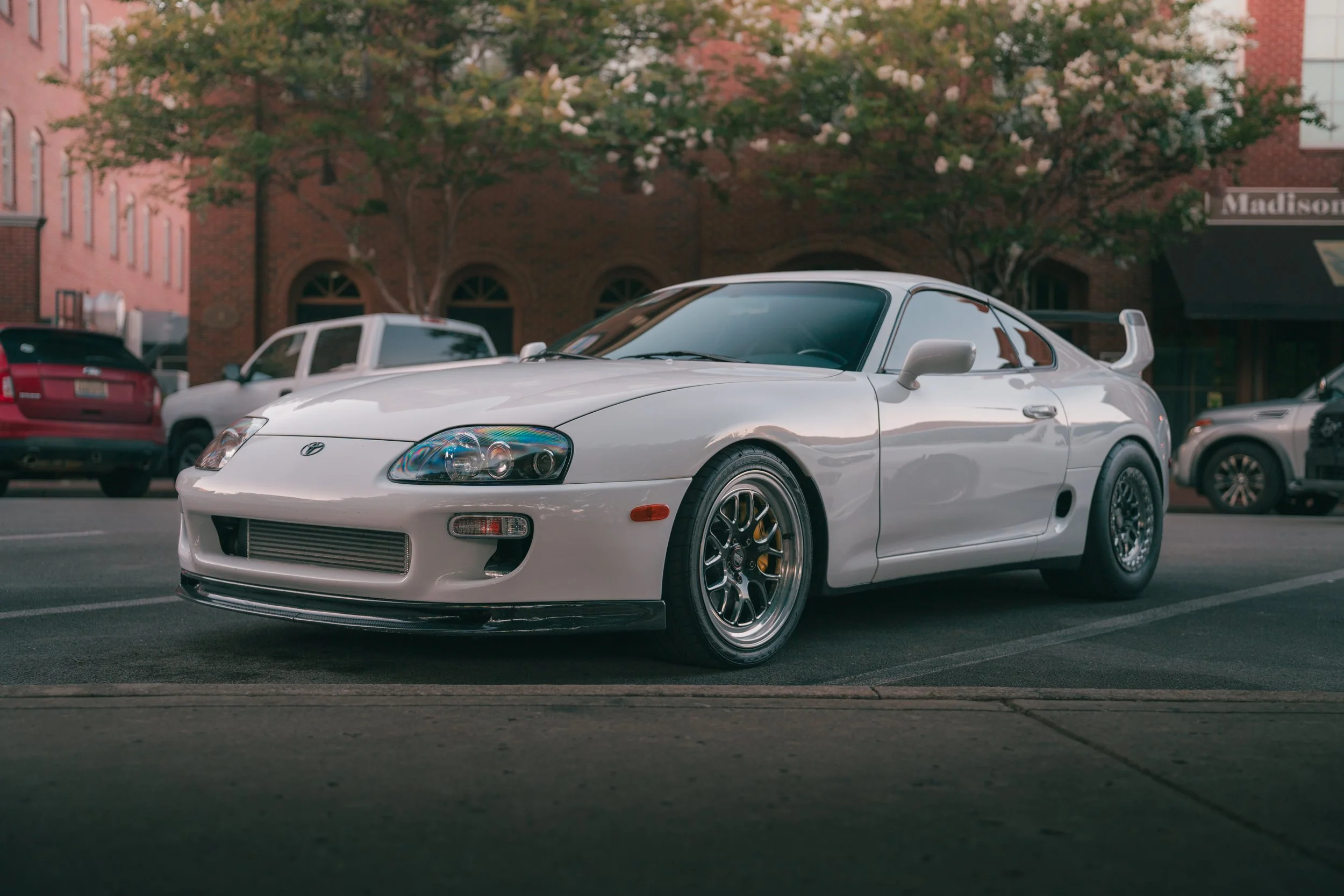 A white Toyota Supra sports car parked on the street during daytime, with other vehicles and brick buildings in the background, and trees with white blossoms overhead.