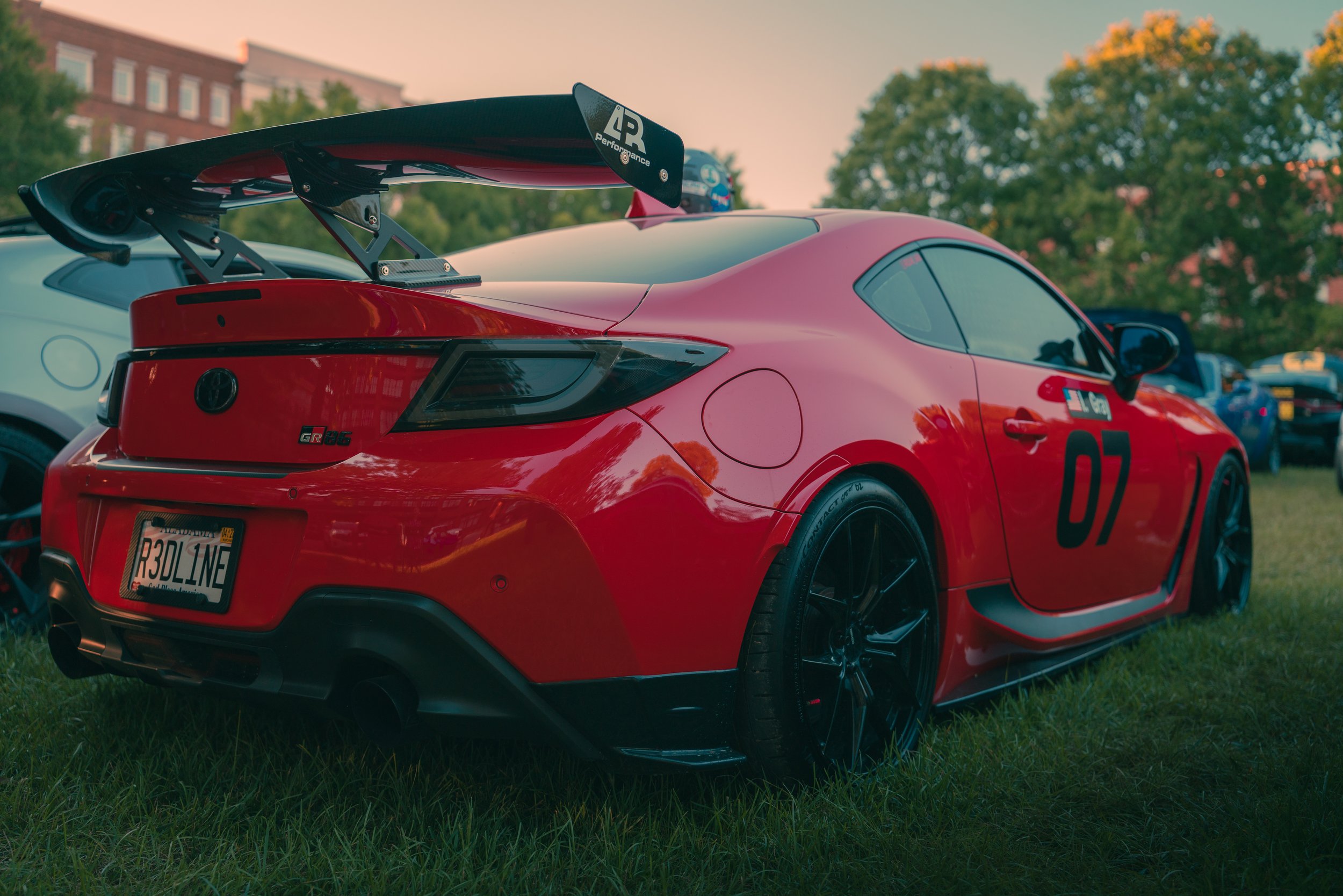 Red sports car with a large rear wing spoiler parked on grass during sunset.