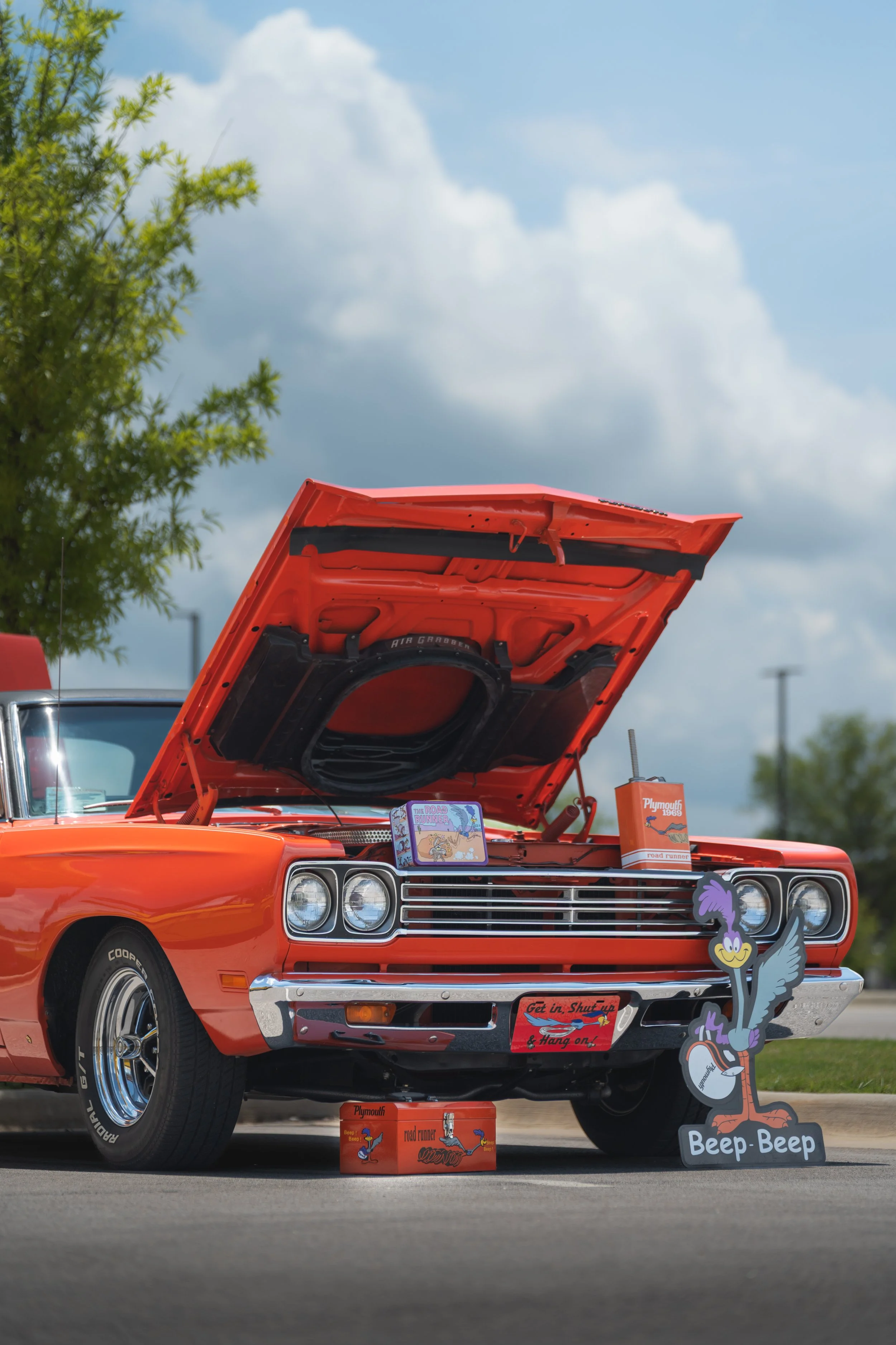 A vintage orange Plymouth car with its hood open, displaying various memorabilia and decorations including a cartoon character cutout with a microphone, a red sign that reads 'Get in, Shut up & Hang on!', and a small orange box labeled 'Road Runner'. The car is parked outdoors under a partly cloudy sky, with a green tree on the left side.