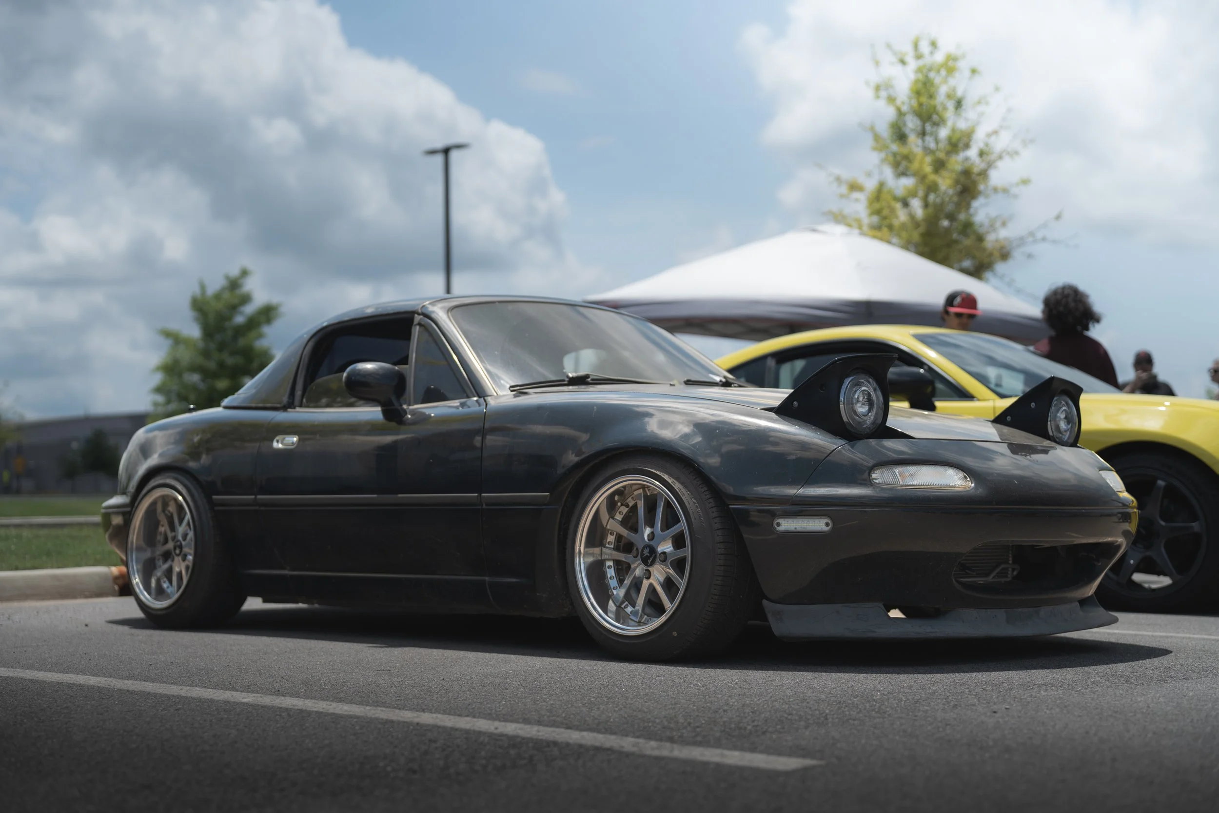 A black Mazda MX-5 Miata with modified wheels and fender flares, parked in a lot during an outdoor car event. Other cars and people are visible in the background under a cloudy sky.