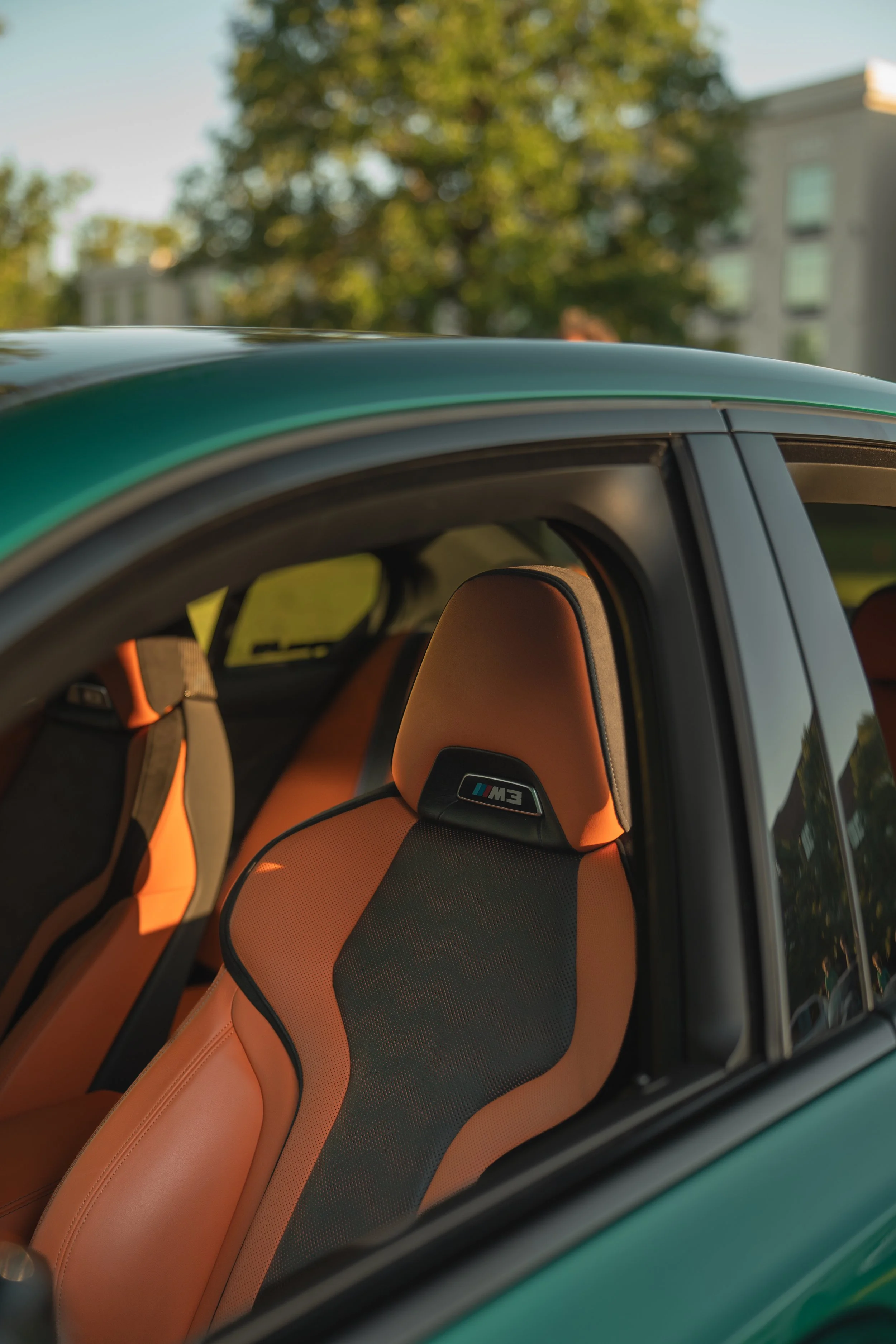 Interior of a car with tan and black seats, including a headrest with an M3 logo, visible through the window with trees and buildings in the background.