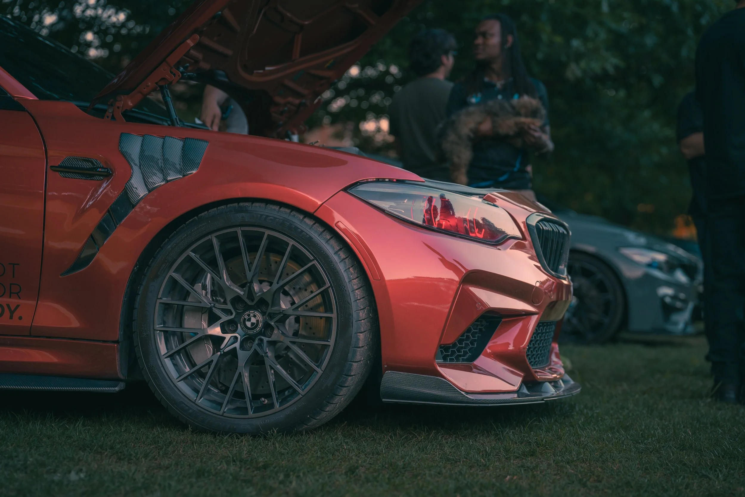 A close-up of a red sports car with its hood open at a car show, with a group of people in the background chatting.