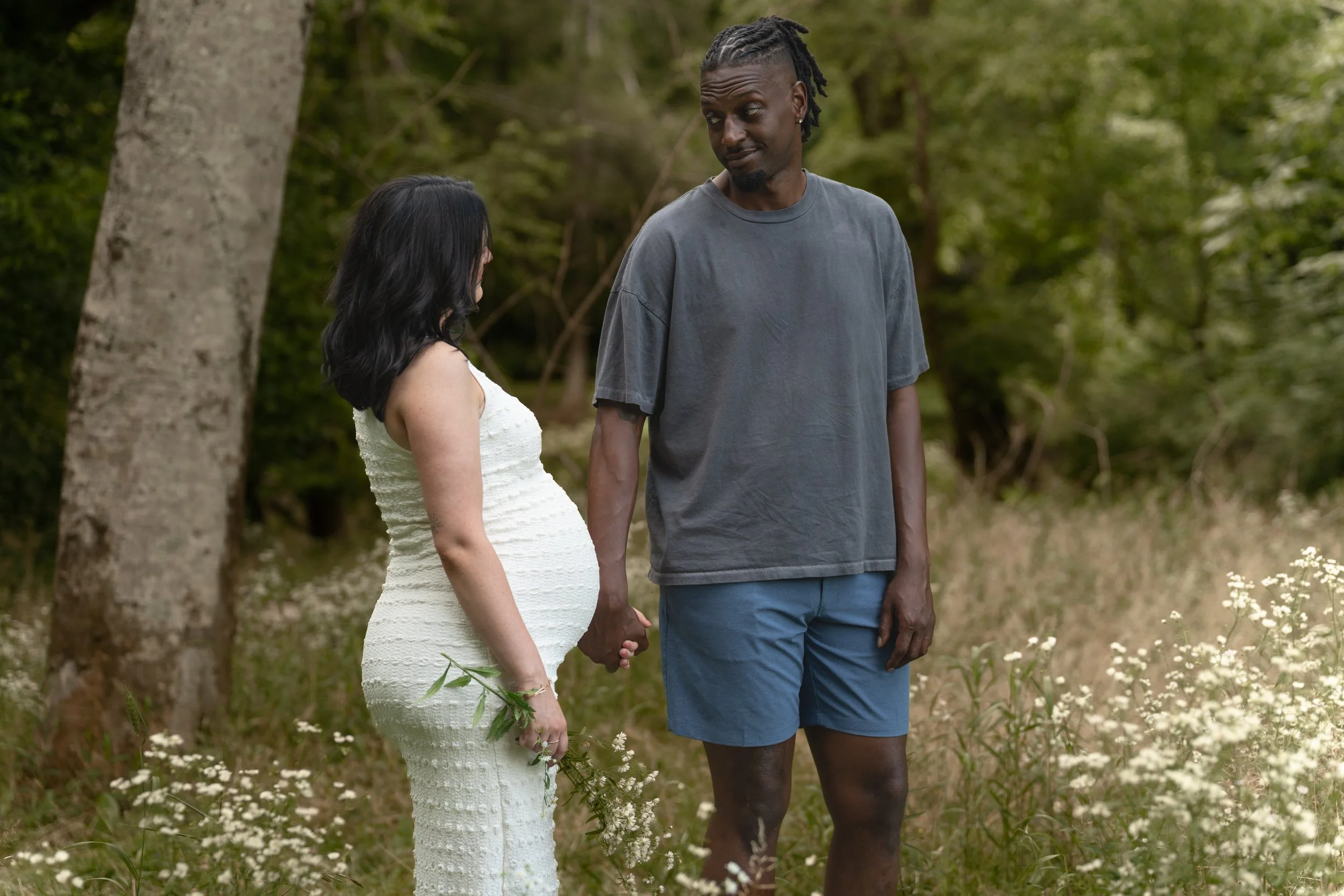 A pregnant woman in a white dress holding a small bouquet of flowers and a man in a blue t-shirt and shorts holding her hand, standing outdoors in a wooded area.
