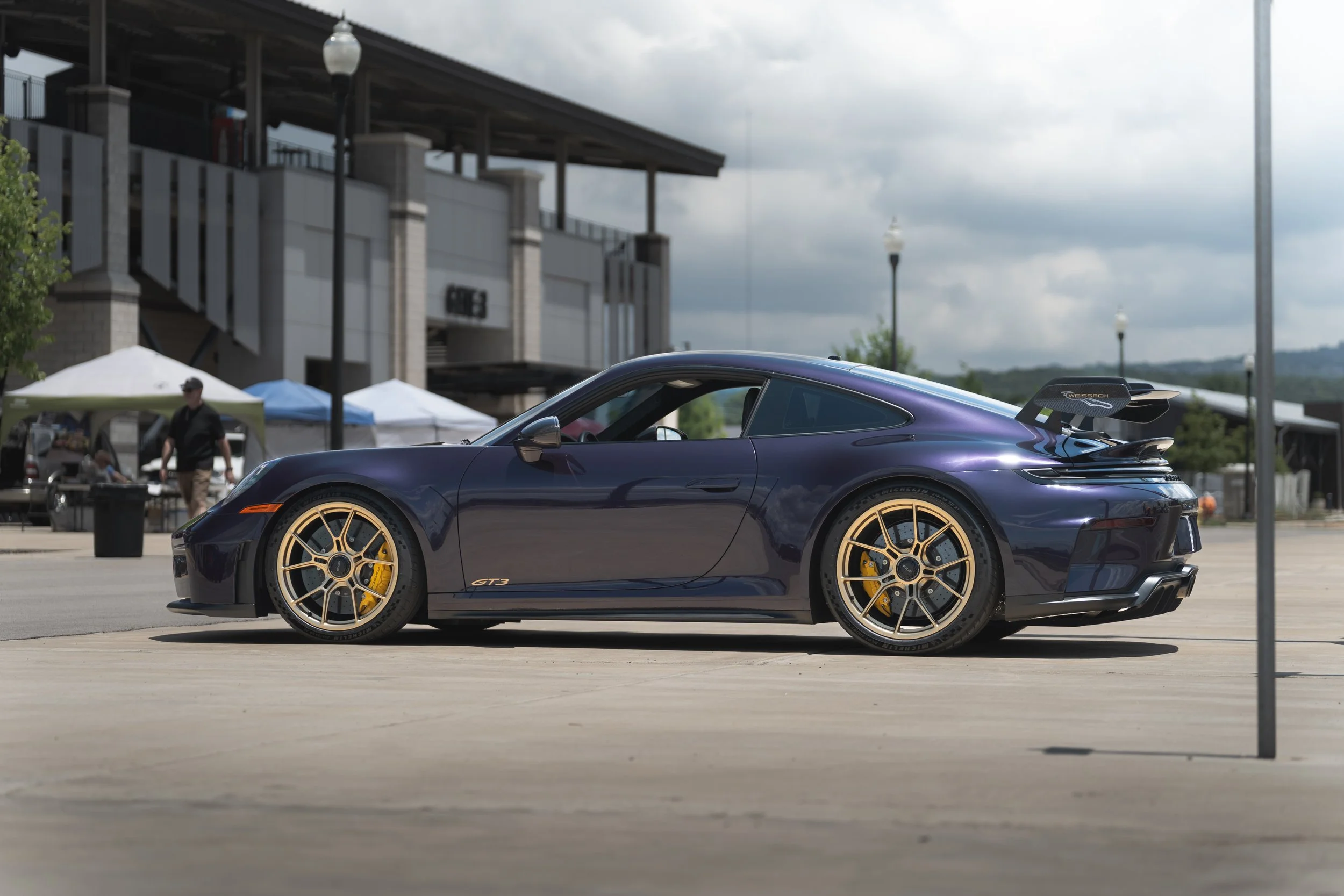 A purple sports car with gold rims parked outside near tents and a building on a cloudy day.