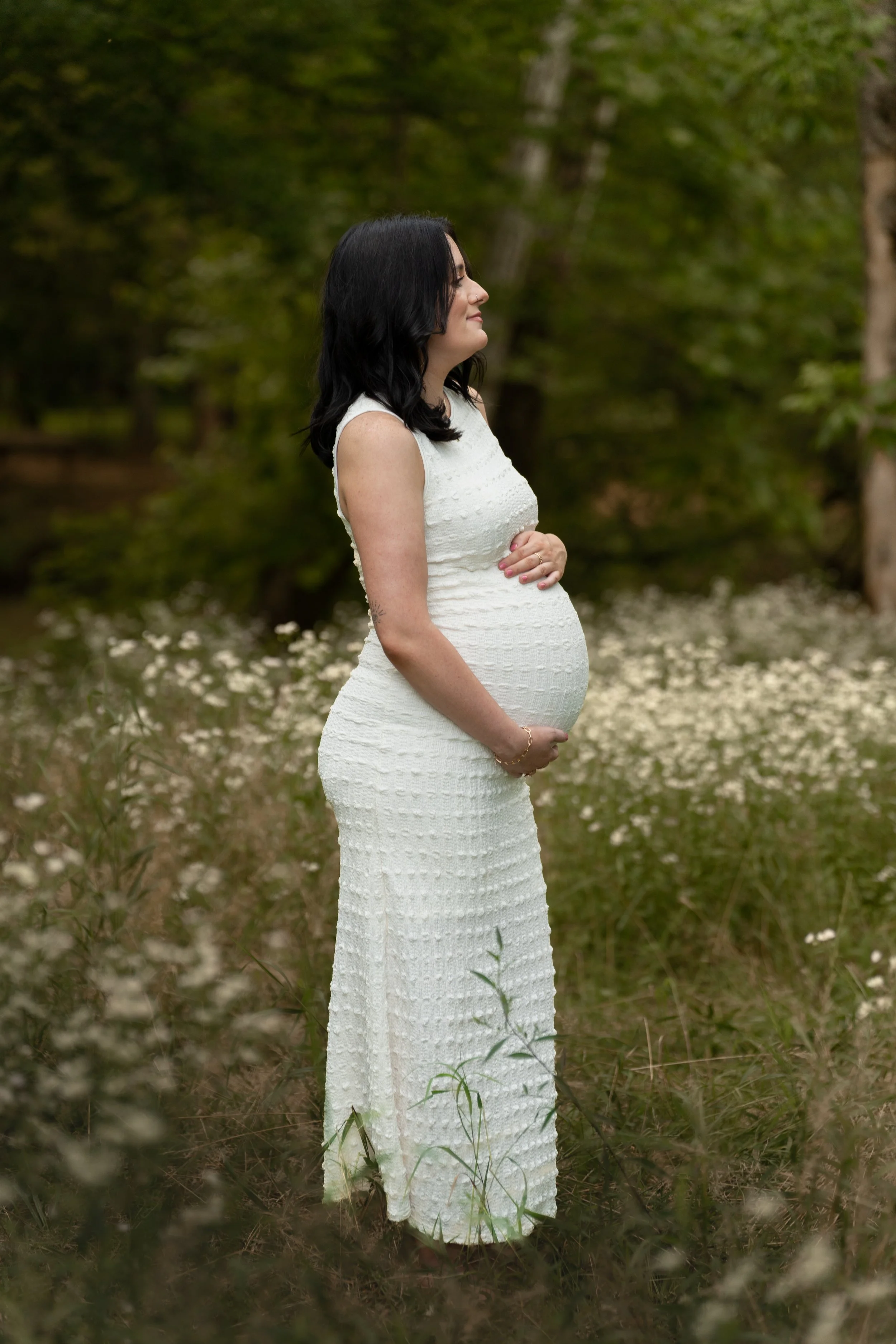 A pregnant woman with dark hair in a white dress standing in a forest with green trees and white flowers.