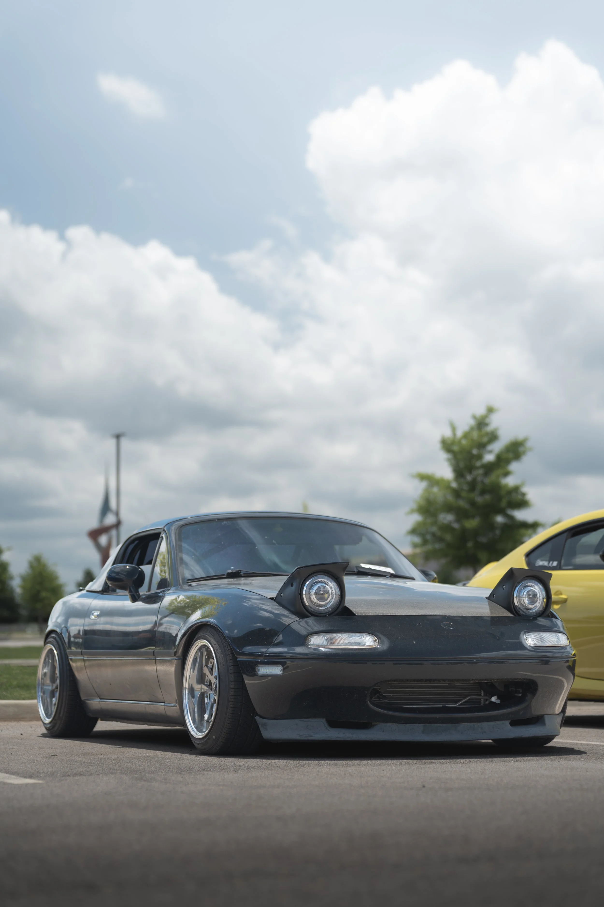 A black sports car with aftermarket modifications parked in a lot, with other cars and an American flag flying in the background under a partly cloudy sky.