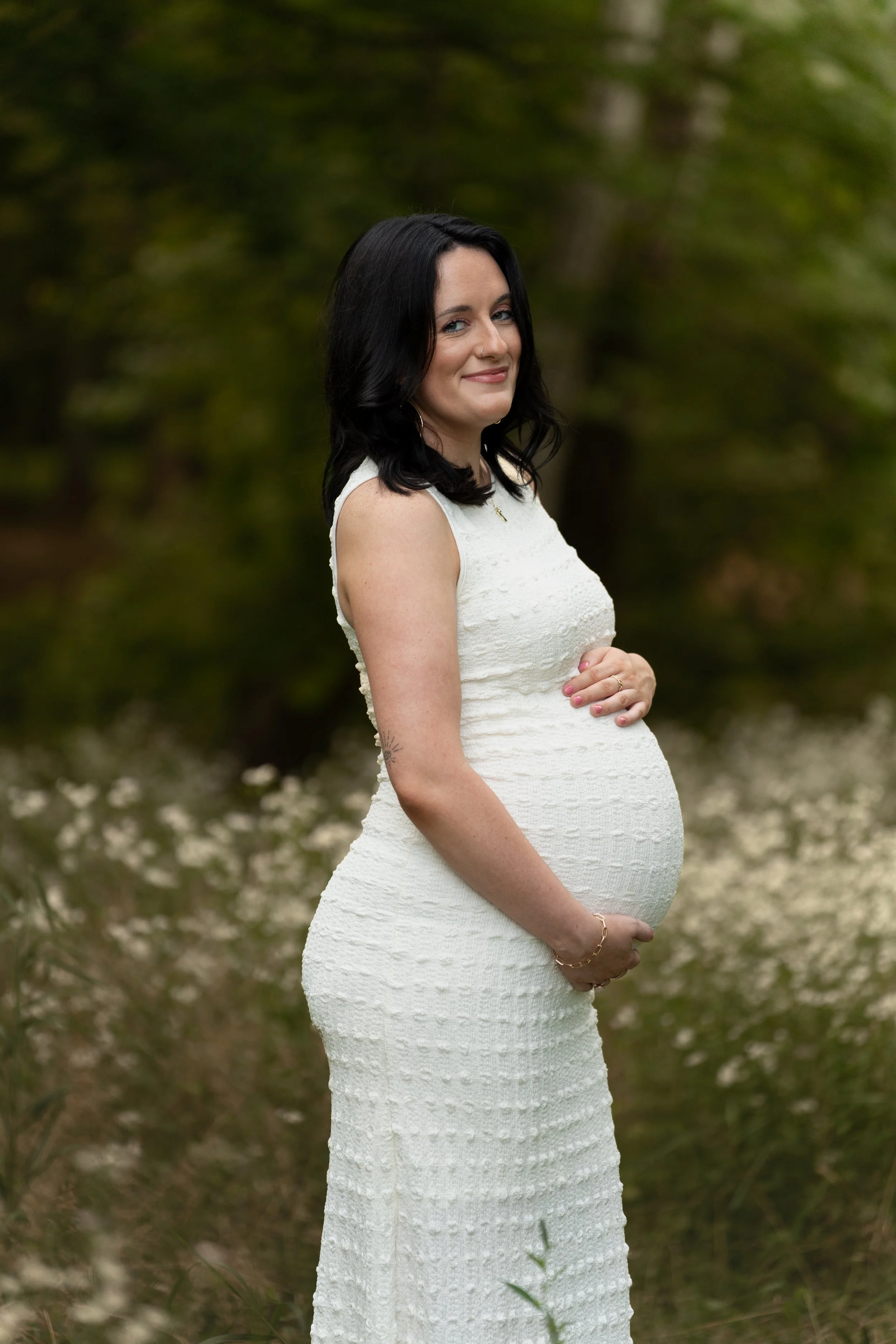 A pregnant woman with black hair in a sleeveless white dress standing outdoors in a wooded area with green foliage and white flowers, smiling at the camera.