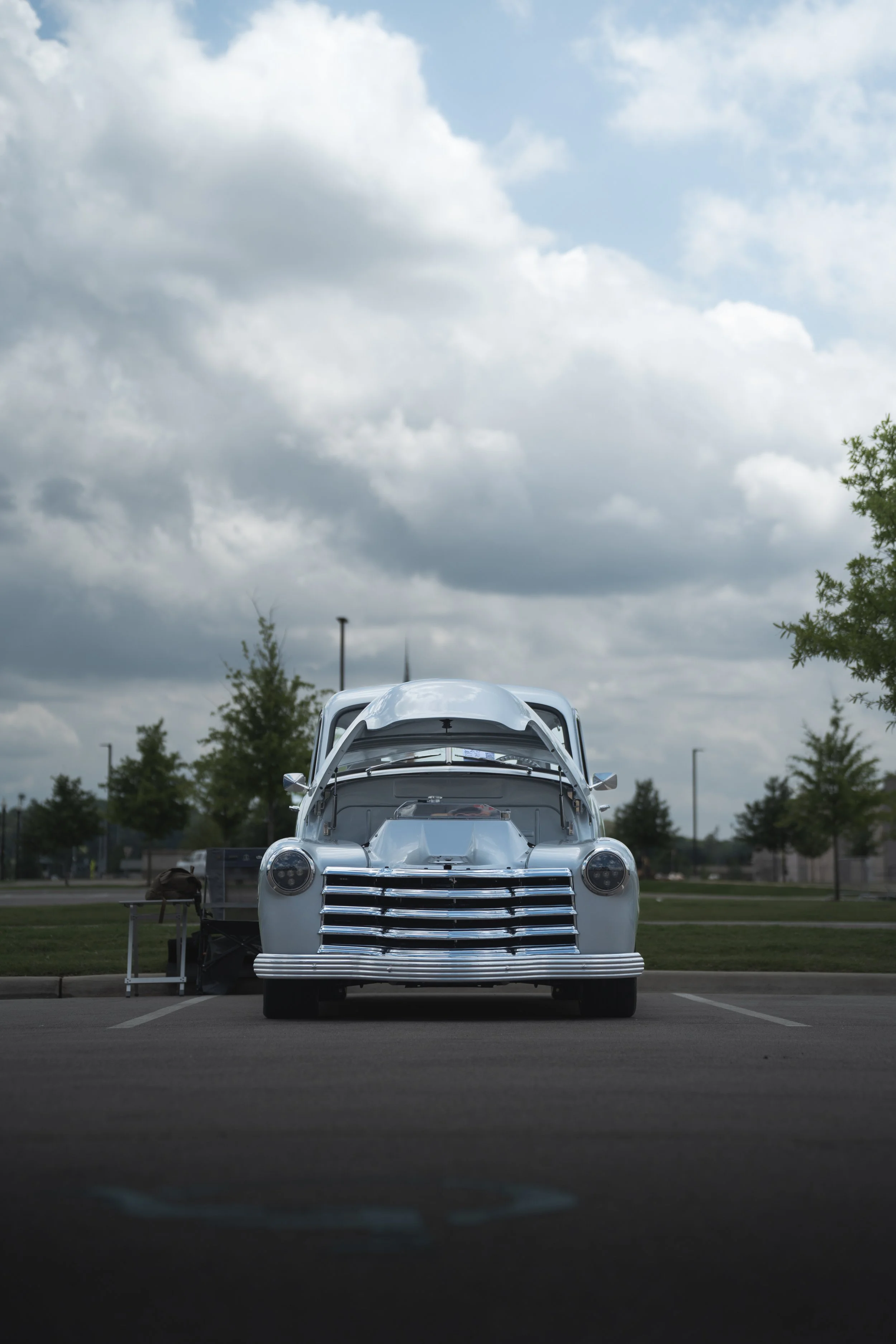 A vintage silver car with its hood open is parked in an empty parking lot, with trees and cloudy sky in the background.