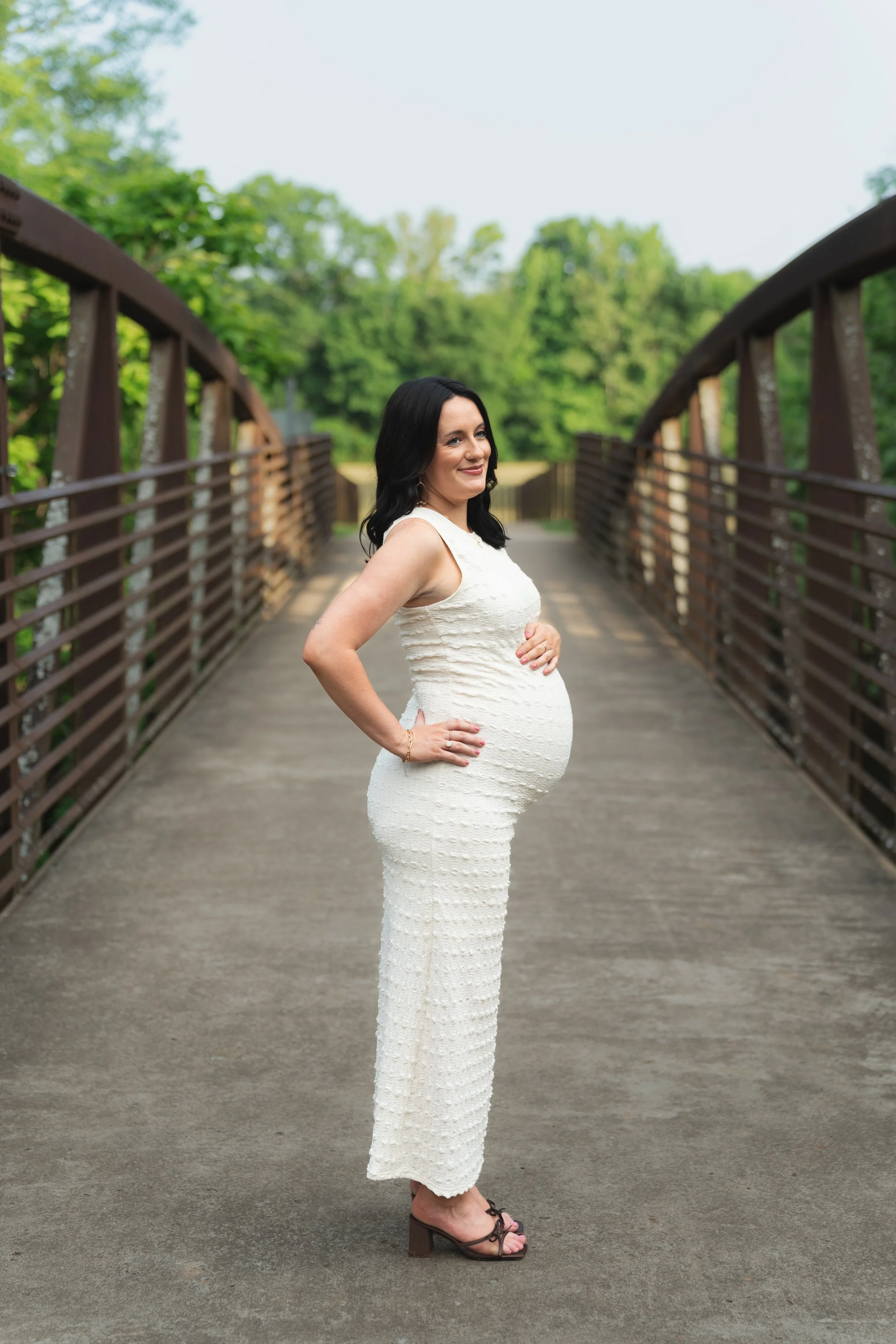 A pregnant woman in a white fitted dress standing on a bridge surrounded by green trees, smiling and looking to her right.