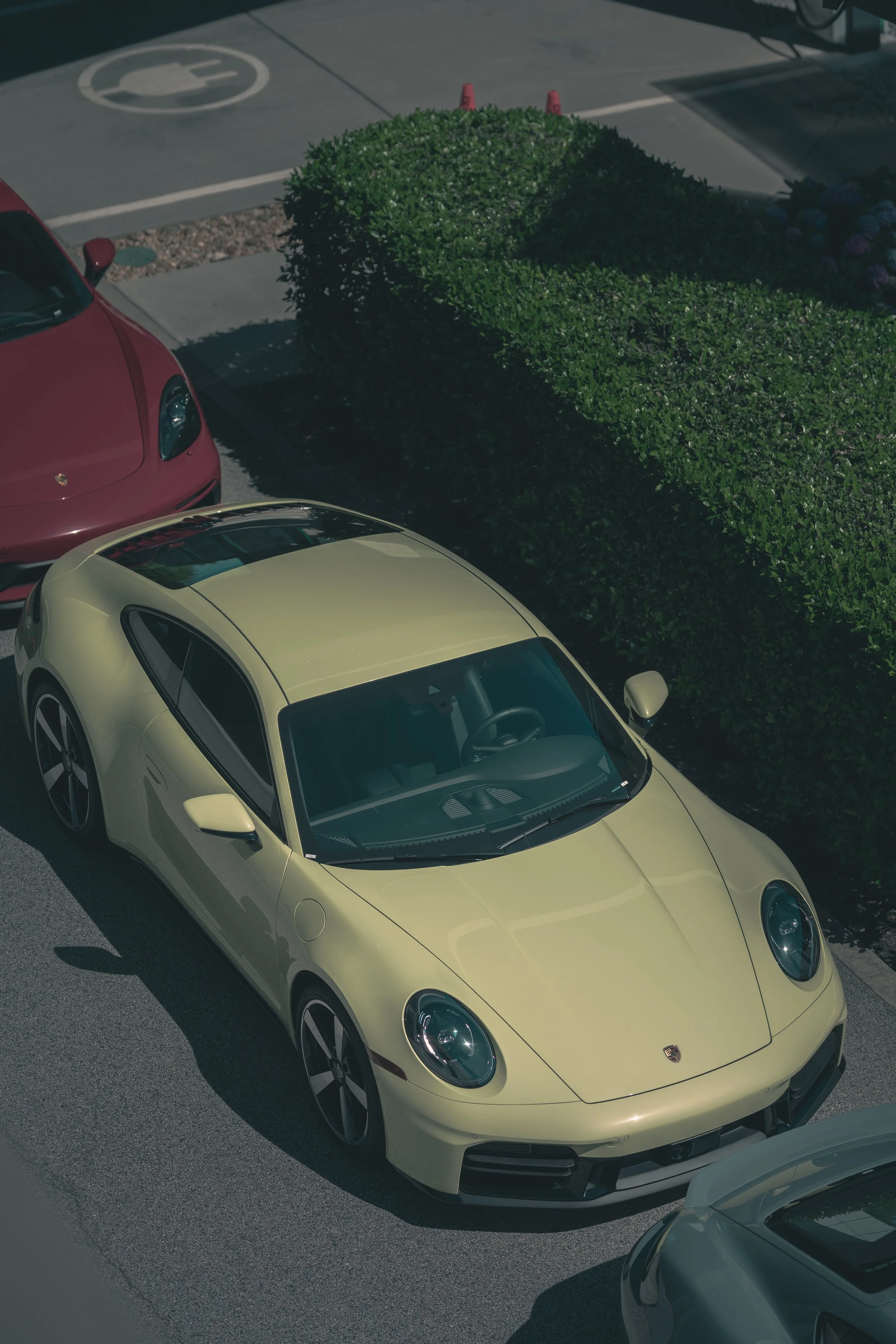 A yellow Porsche parked in a lot next to a hedge, with parts of other vehicles visible nearby. The Porsche has a sleek design with a black roof and is in sunlight, casting a shadow on the ground.