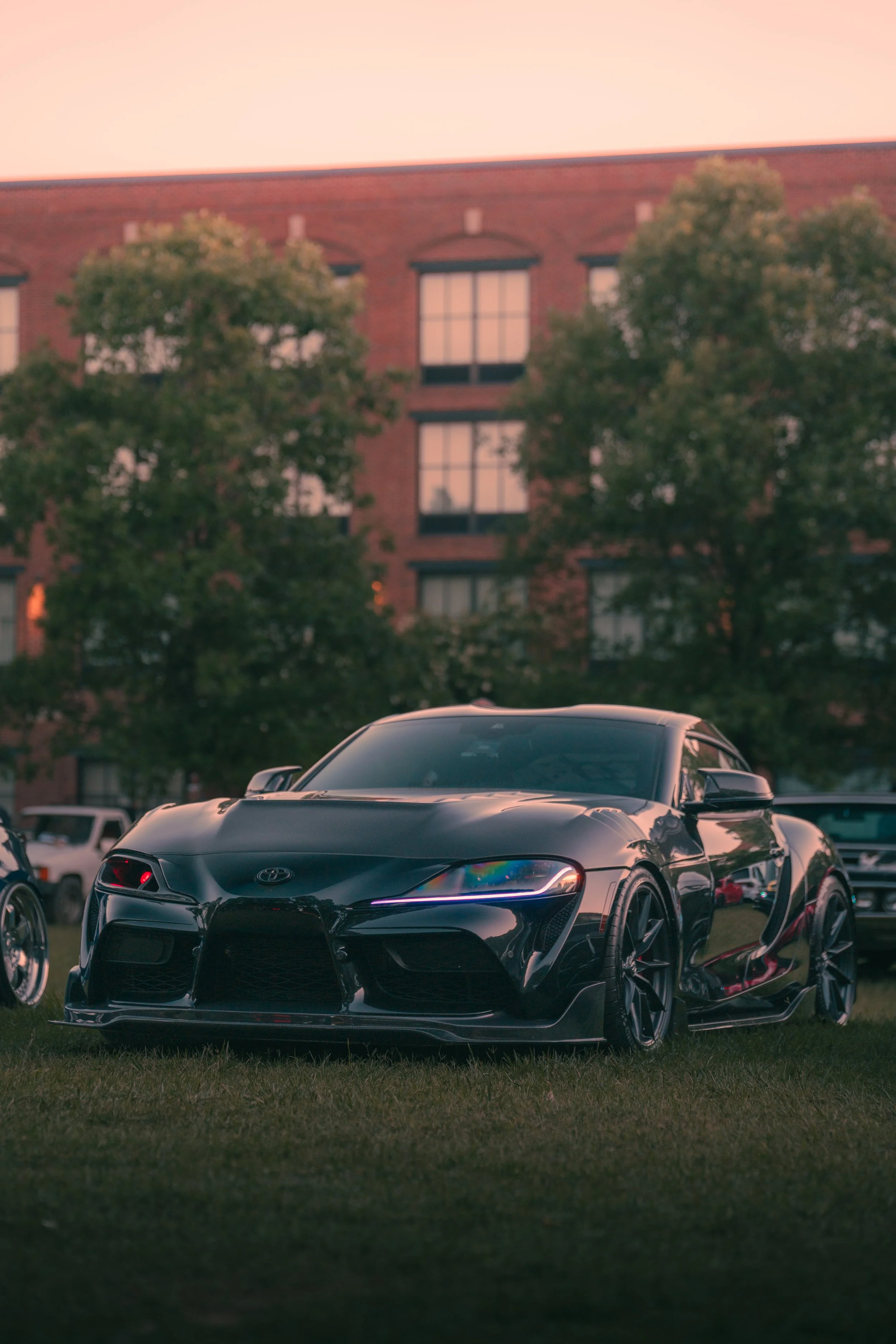 A black sports car parked on grass with a brick building and trees in the background during sunset.