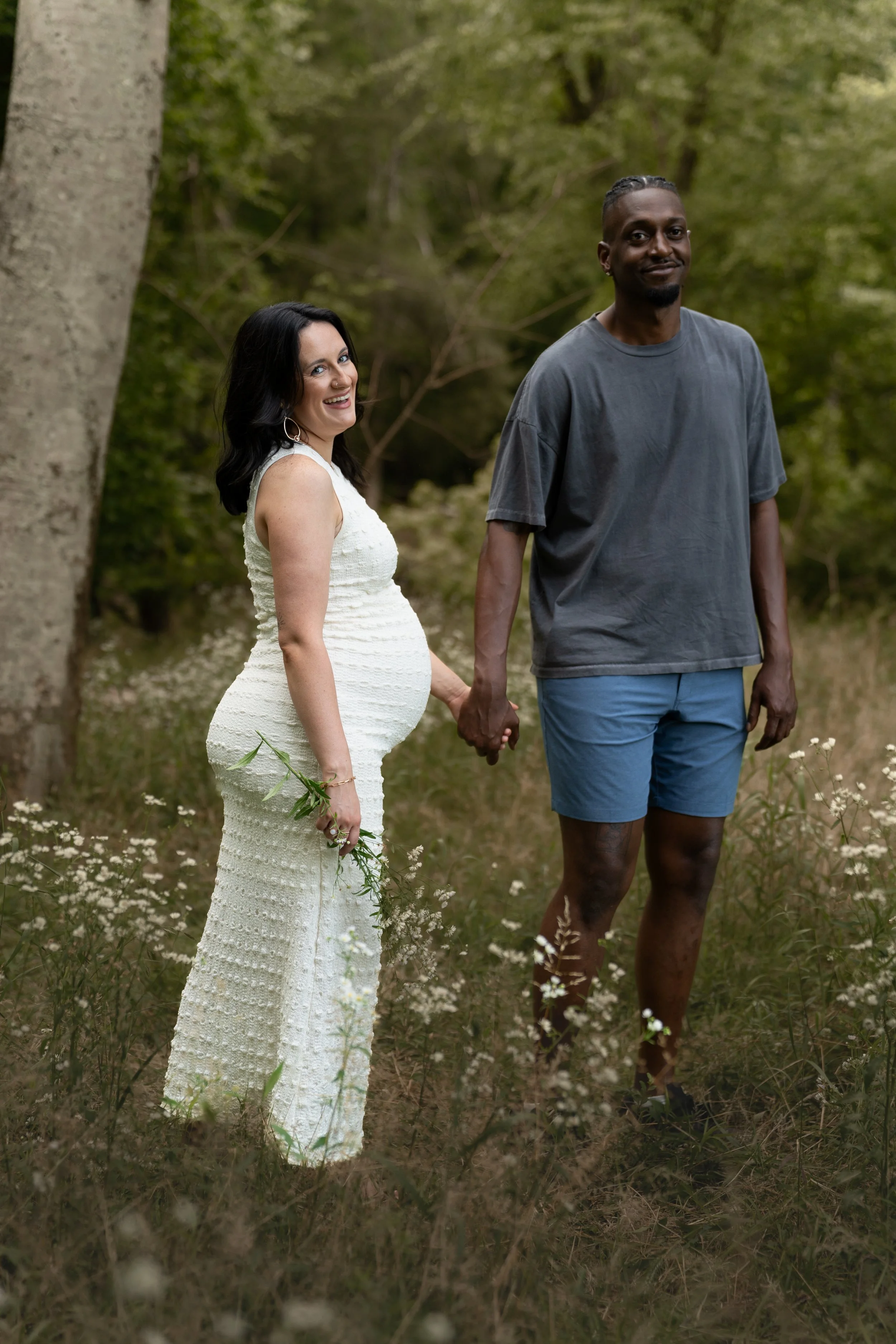 A smiling woman in a white dress holding a small branch, and a man in a gray t-shirt and blue shorts holding her hand, standing outdoors in a forested area with trees and white flowers.