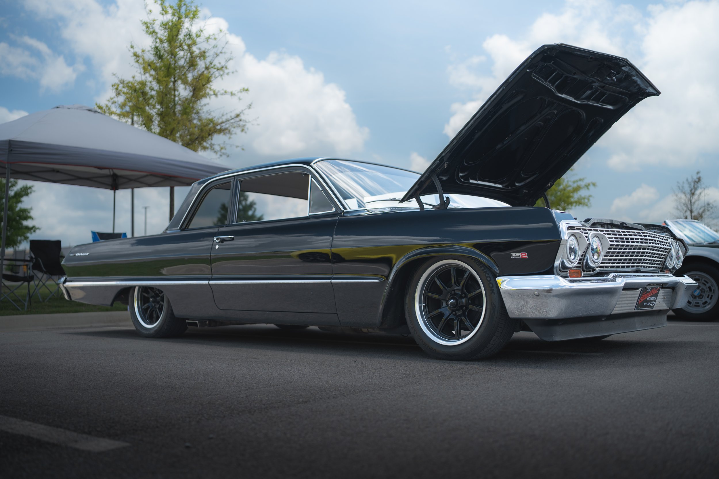 A classic black Buick muscle car with its hood open, parked outdoors at a car show on a sunny day with clouds and trees in the background.