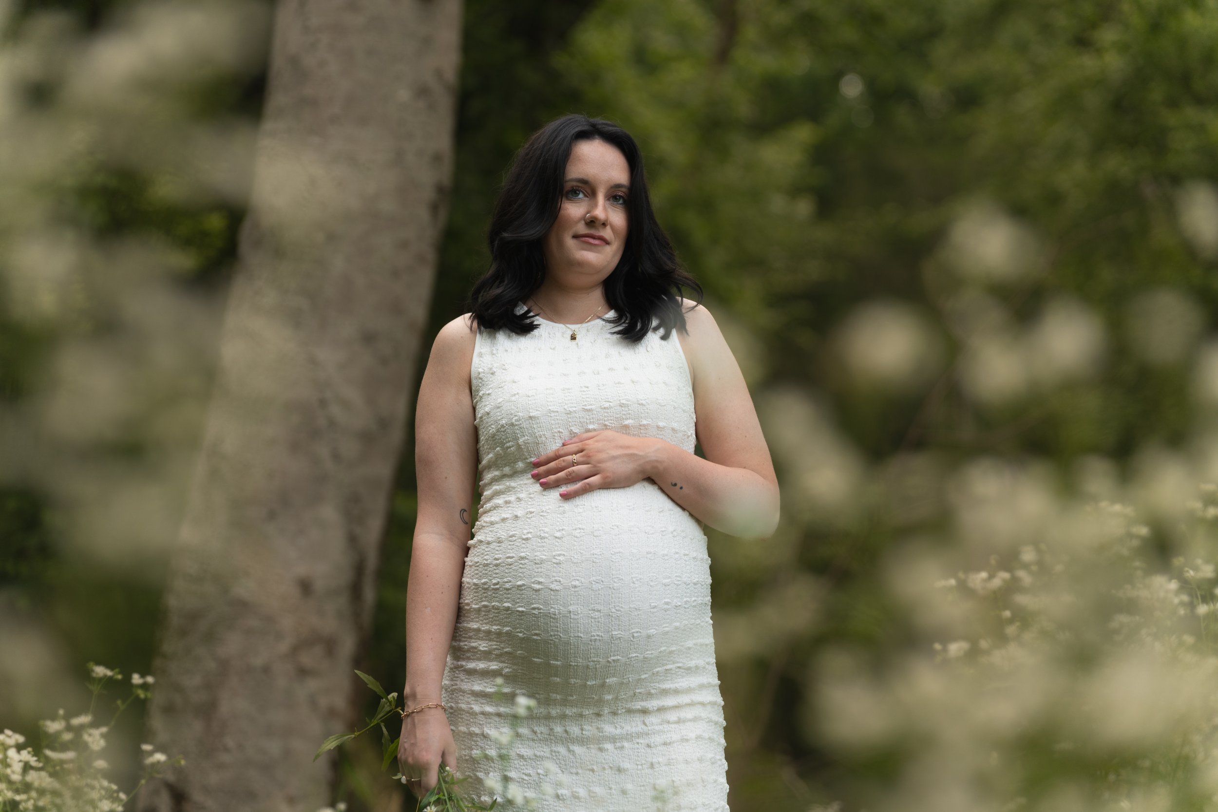 A woman with black hair in a white sleeveless dress standing outdoors near trees, with a gentle expression and her hand on her stomach.