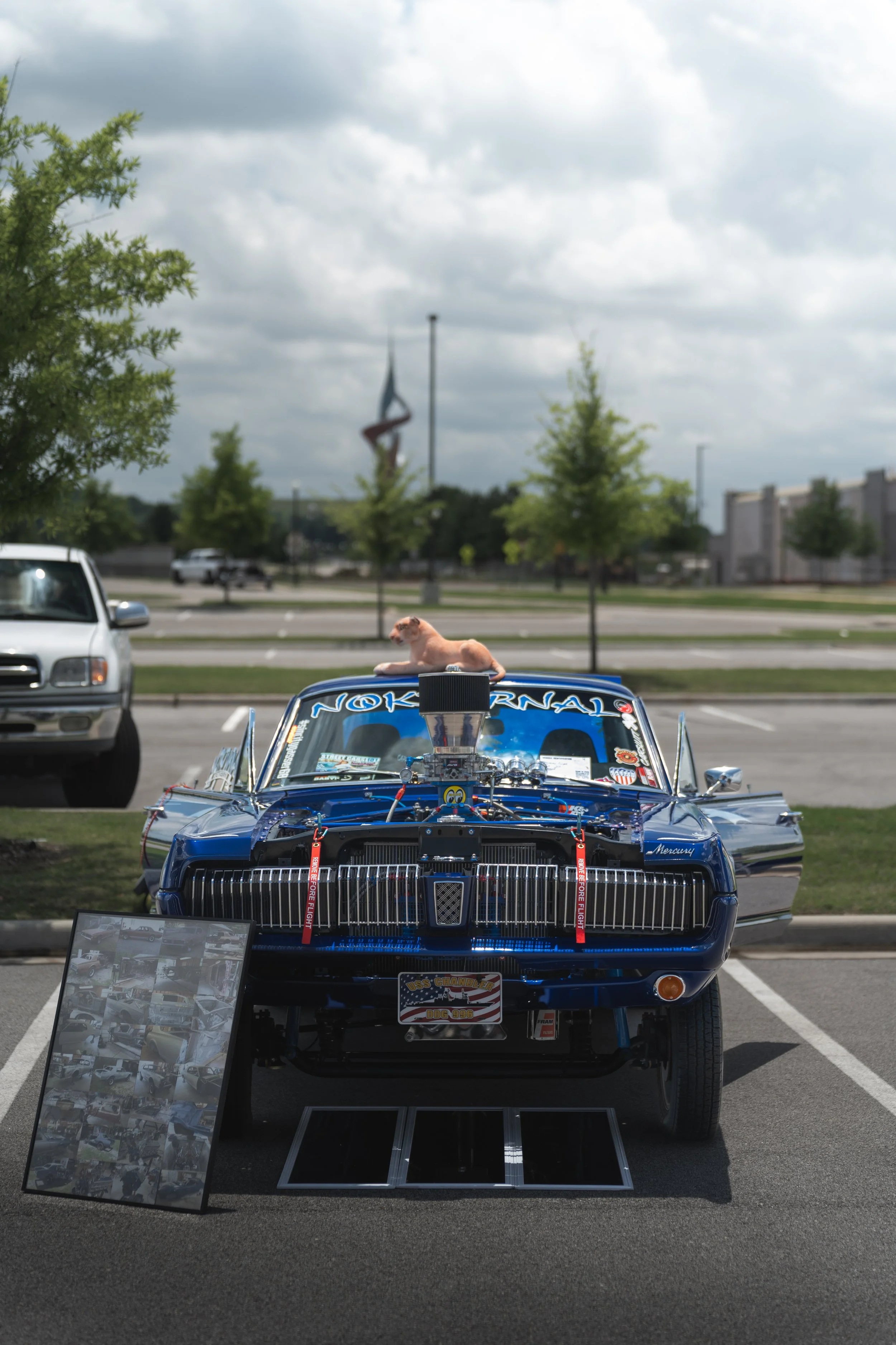 A blue drag racing car displayed in a parking lot with its hood open, a small stuffed dog lying on top, and a photo collage on a stand nearby. The car has stickers and parts indicating it is modified for racing.