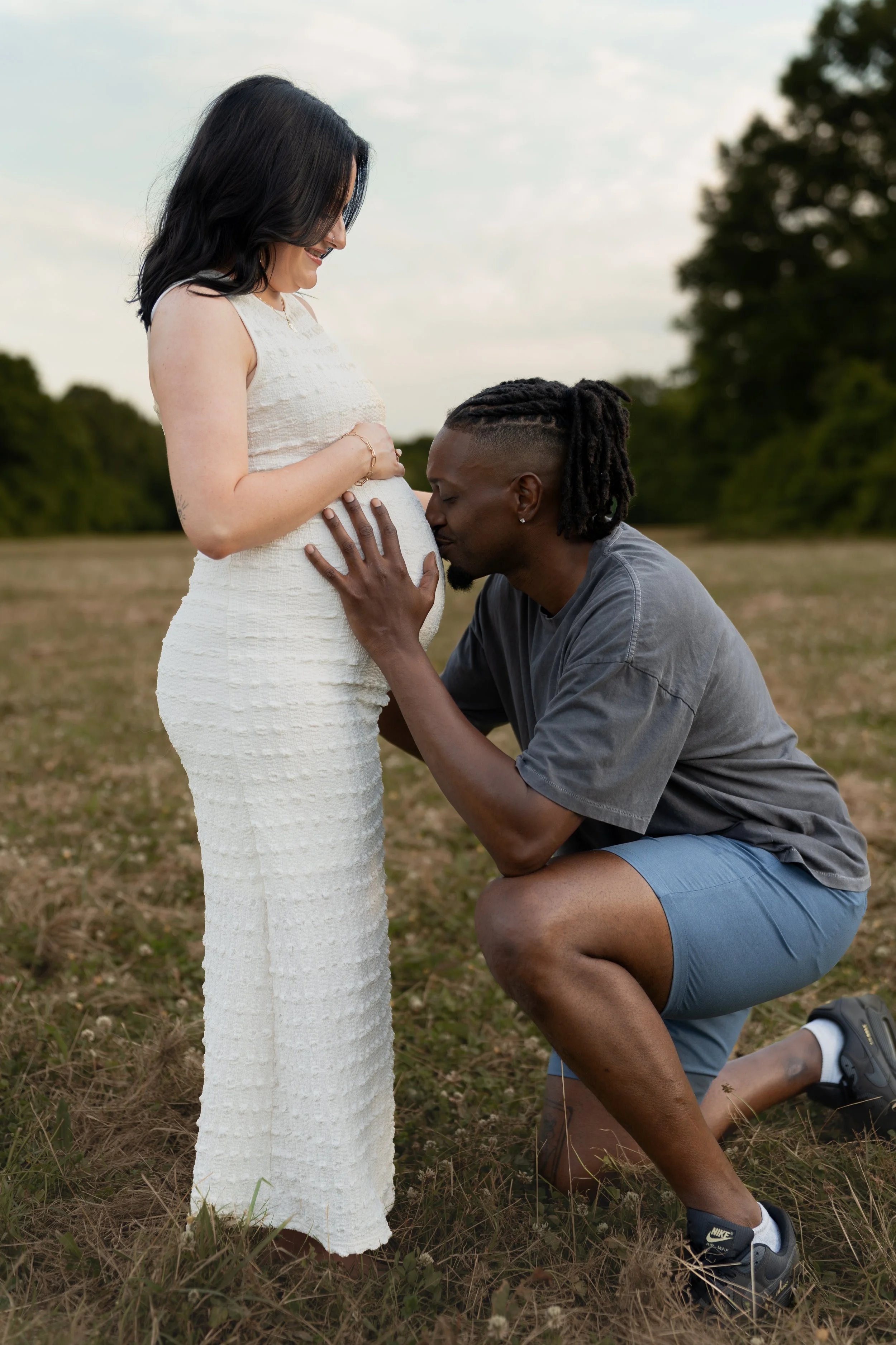 A man kneeling on the ground with one knee, holding a pregnant woman's belly and kissing it, outdoors in a field with trees in the background. The woman is standing in a white dress, smiling, with her hand on her belly.