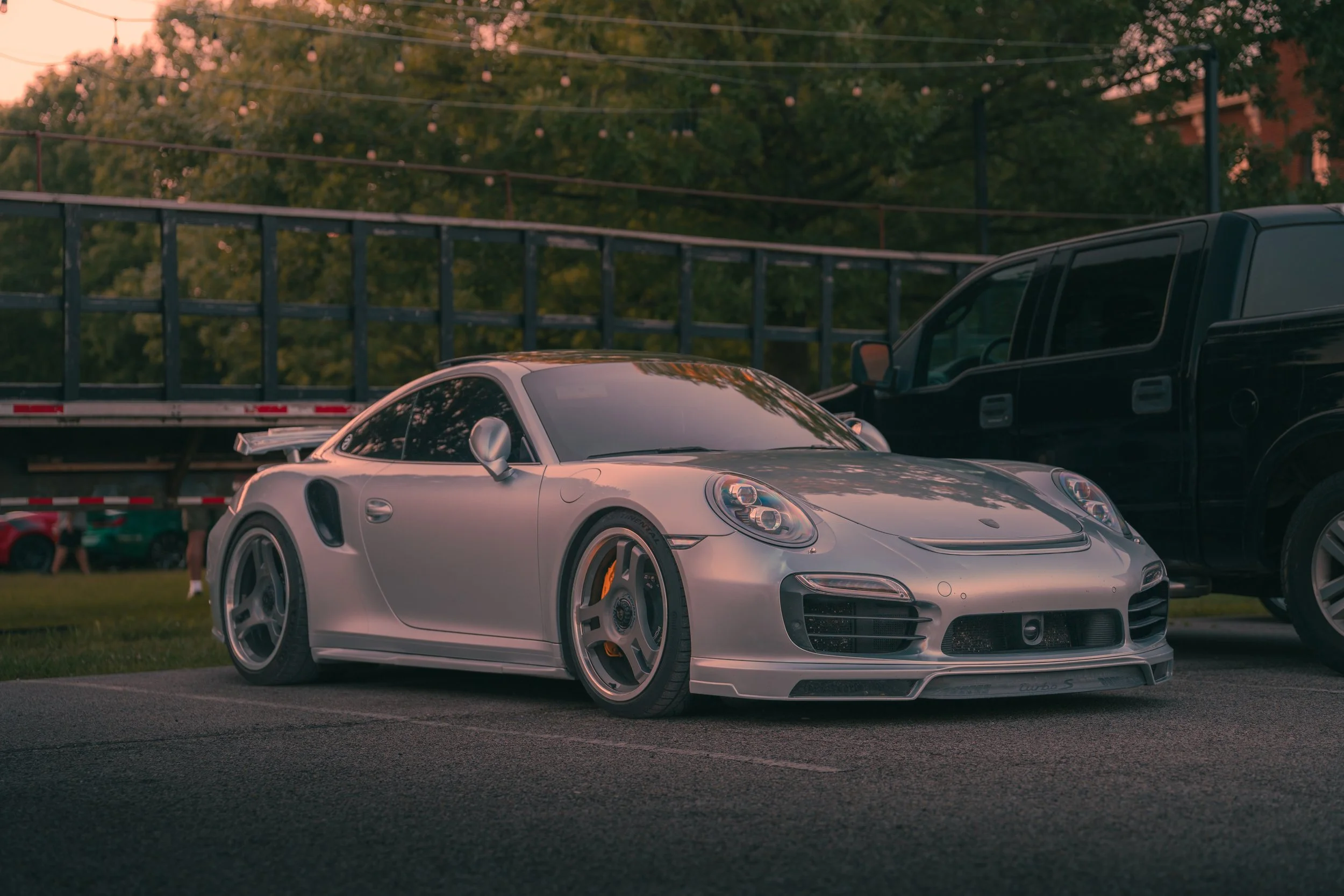 A silver sports car parked in a lot during sunset with a black vehicle next to it, trees, and string lights overhead.