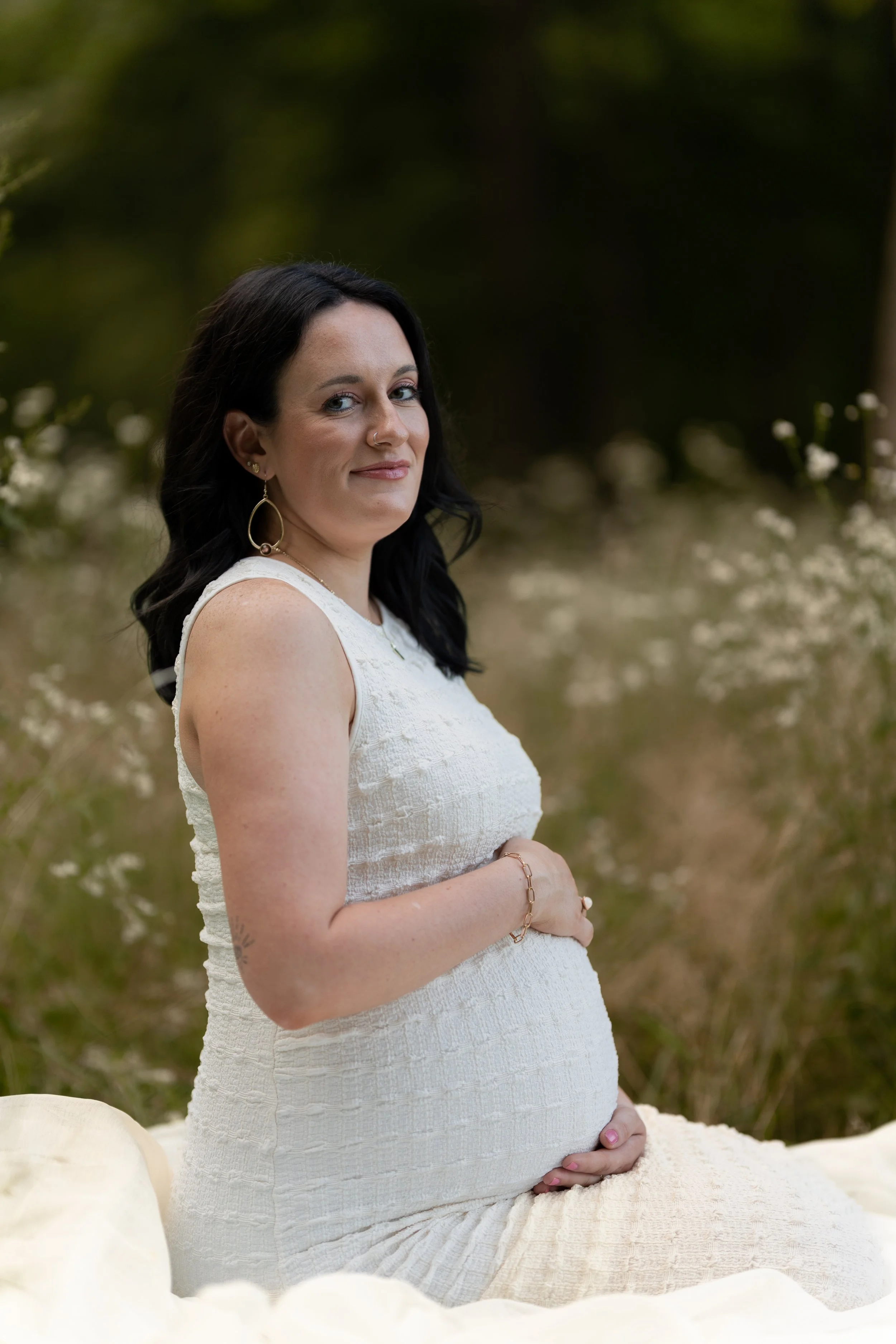 Pregnant woman with dark hair and blue eyes sitting outdoors on a cream-colored blanket in a natural setting with blurred greenery background.