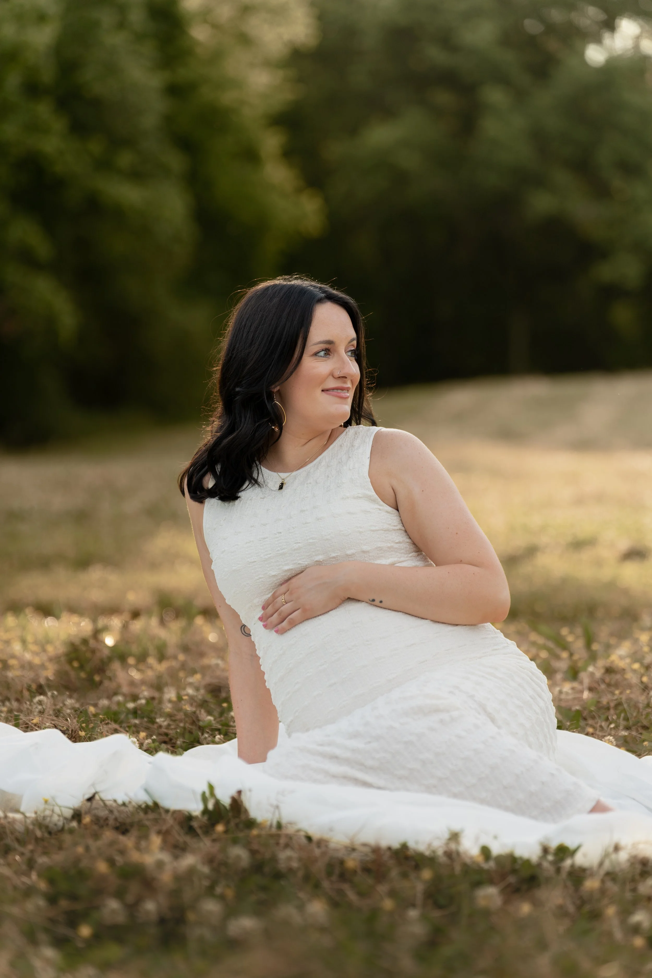 A woman wearing a white dress sitting outdoors on the grass with trees in the background.