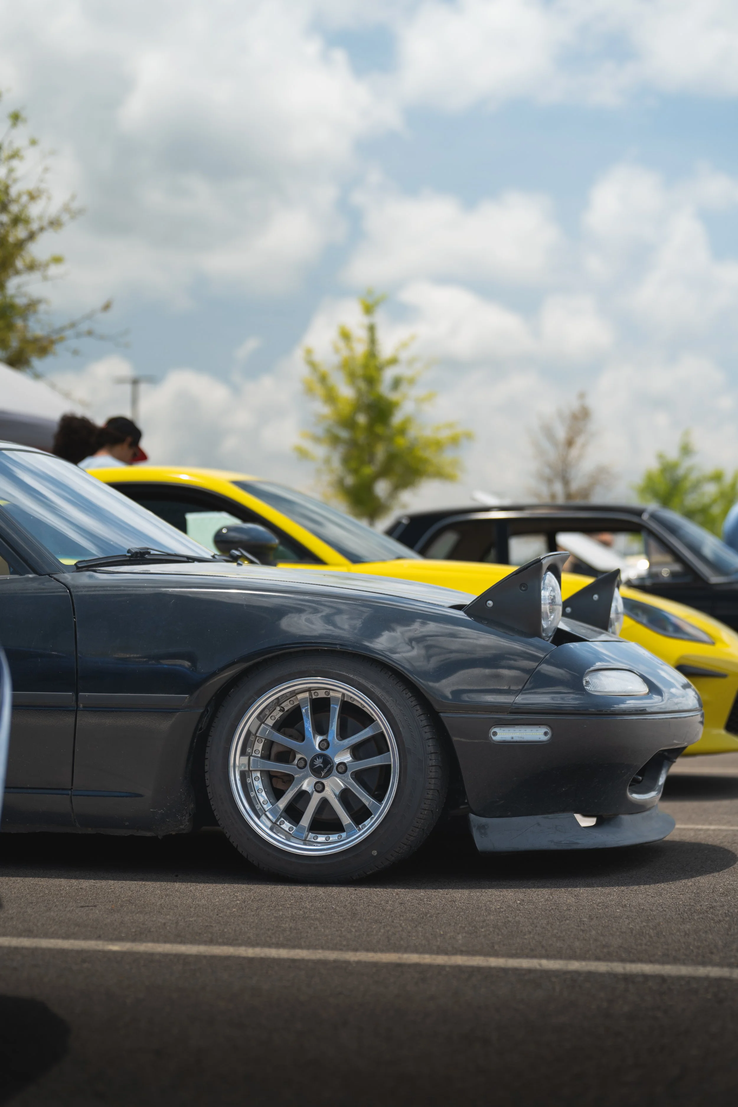 A lineup of sports cars parked outdoors on a cloudy day, with a focus on a black car in the foreground, followed by a yellow car and other vehicles in the background.