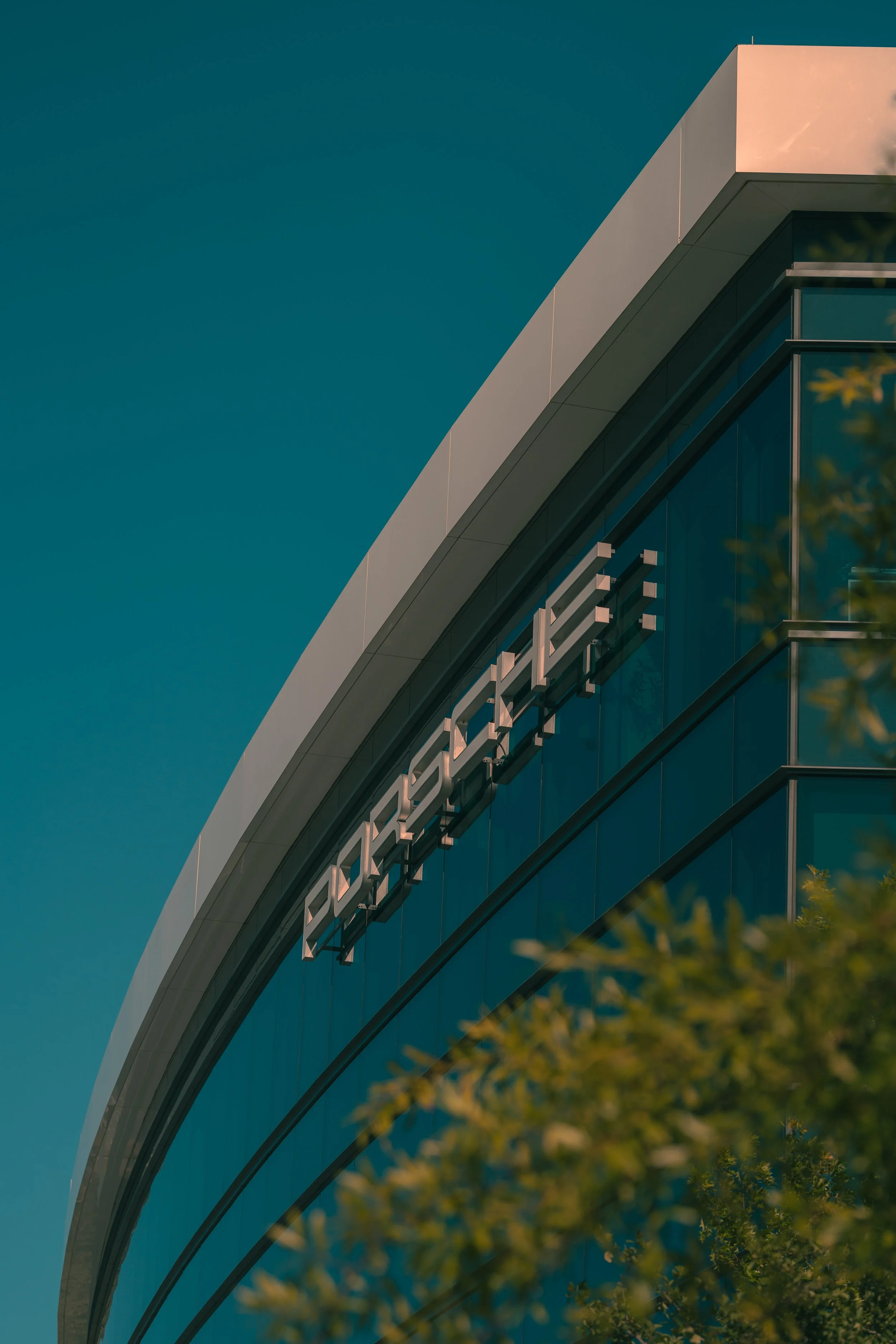 Close-up of a modern building with glass walls and a sign that reads 'EMERGENCY' in large letters, partially obscured by tree leaves in the foreground.