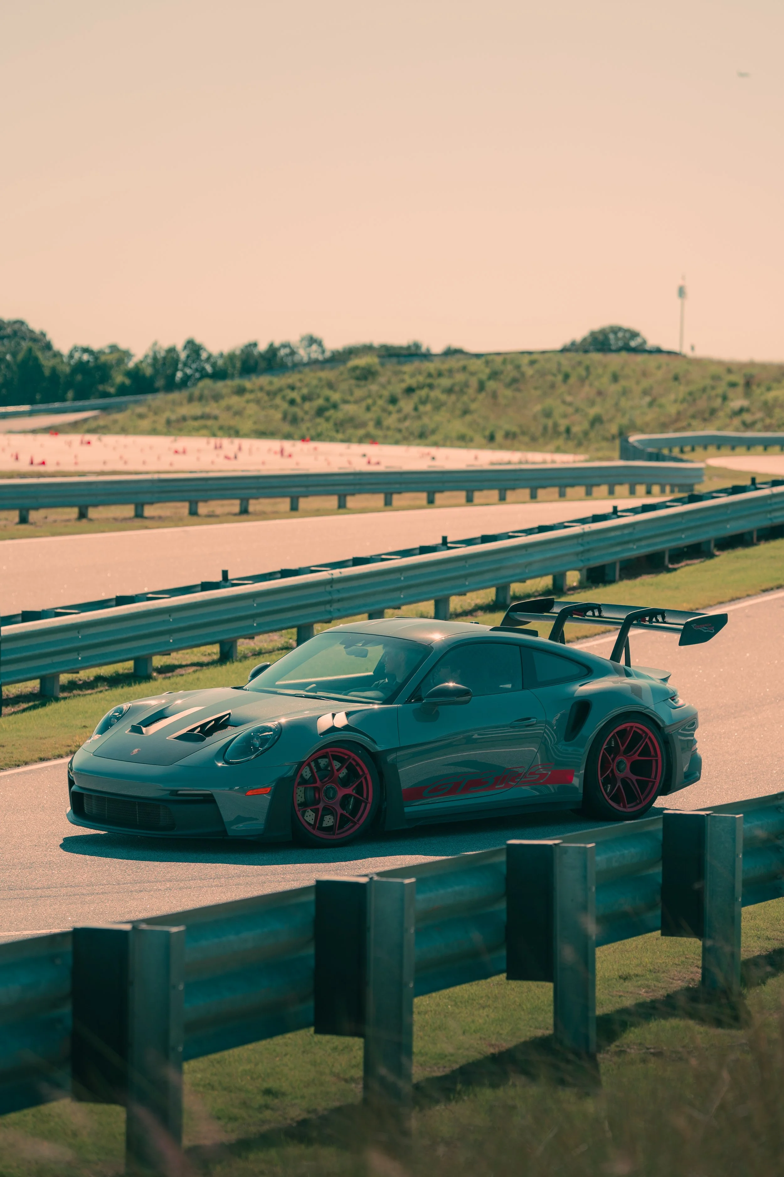 A black sports car with red wheels and racing decals on a race track, with safety barriers and a grassy hill in the background.
