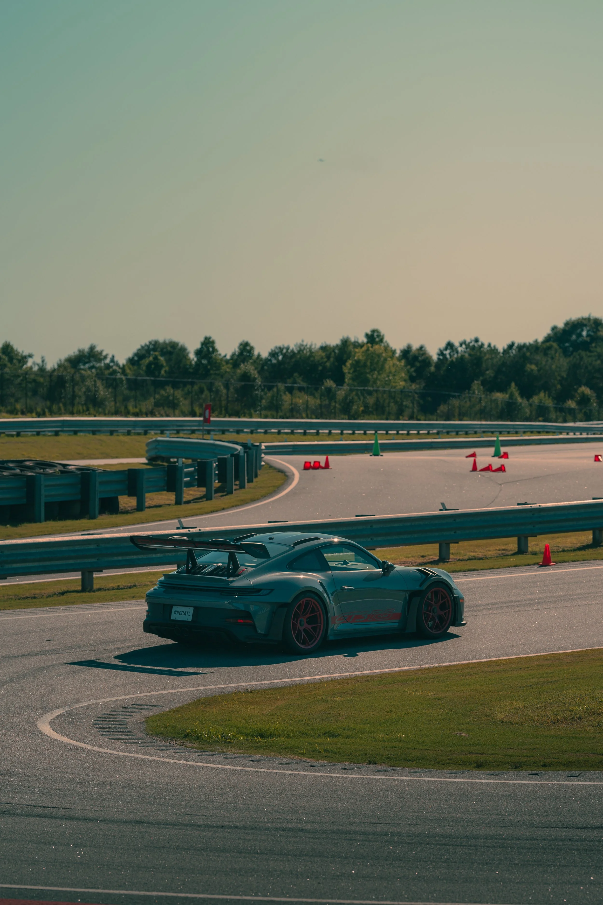 A gray sports car with red accents and a rear wing, parked on the side of a race track next to a guardrail, with orange cones and a curved section of the track in the background, under a mostly clear sky with a faint presence of a small plane.