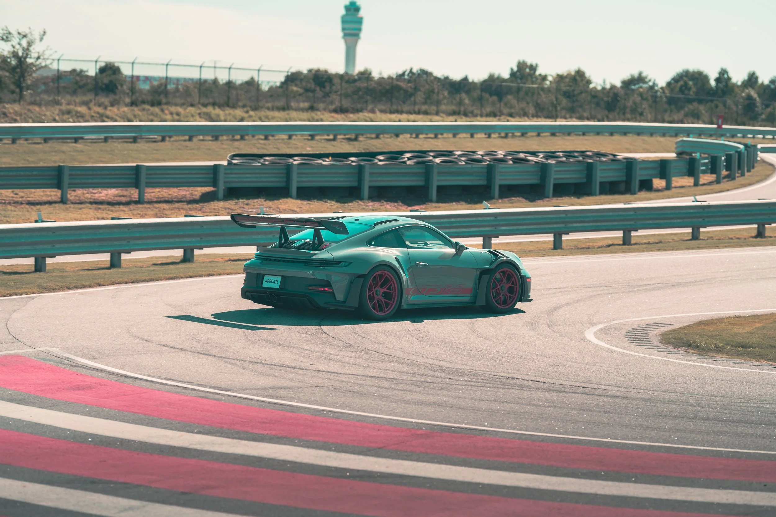 A teal-colored sports car with a large rear spoiler driving on a race track with tire barriers and guardrails, with a control tower in the background under a cloudy sky.