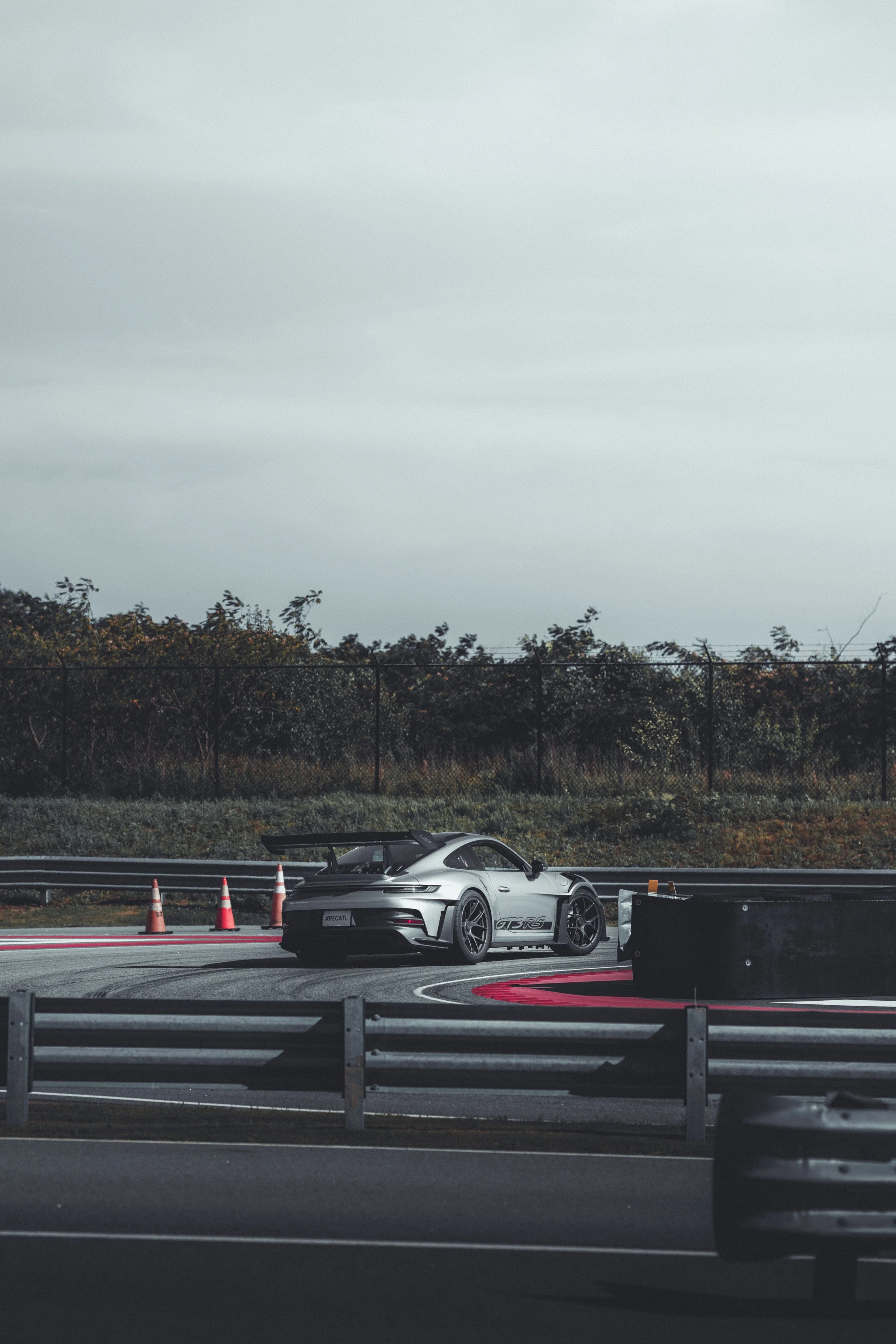 A silver sports car on a race track, with cones and barriers, and a forest and cloudy sky in the background.