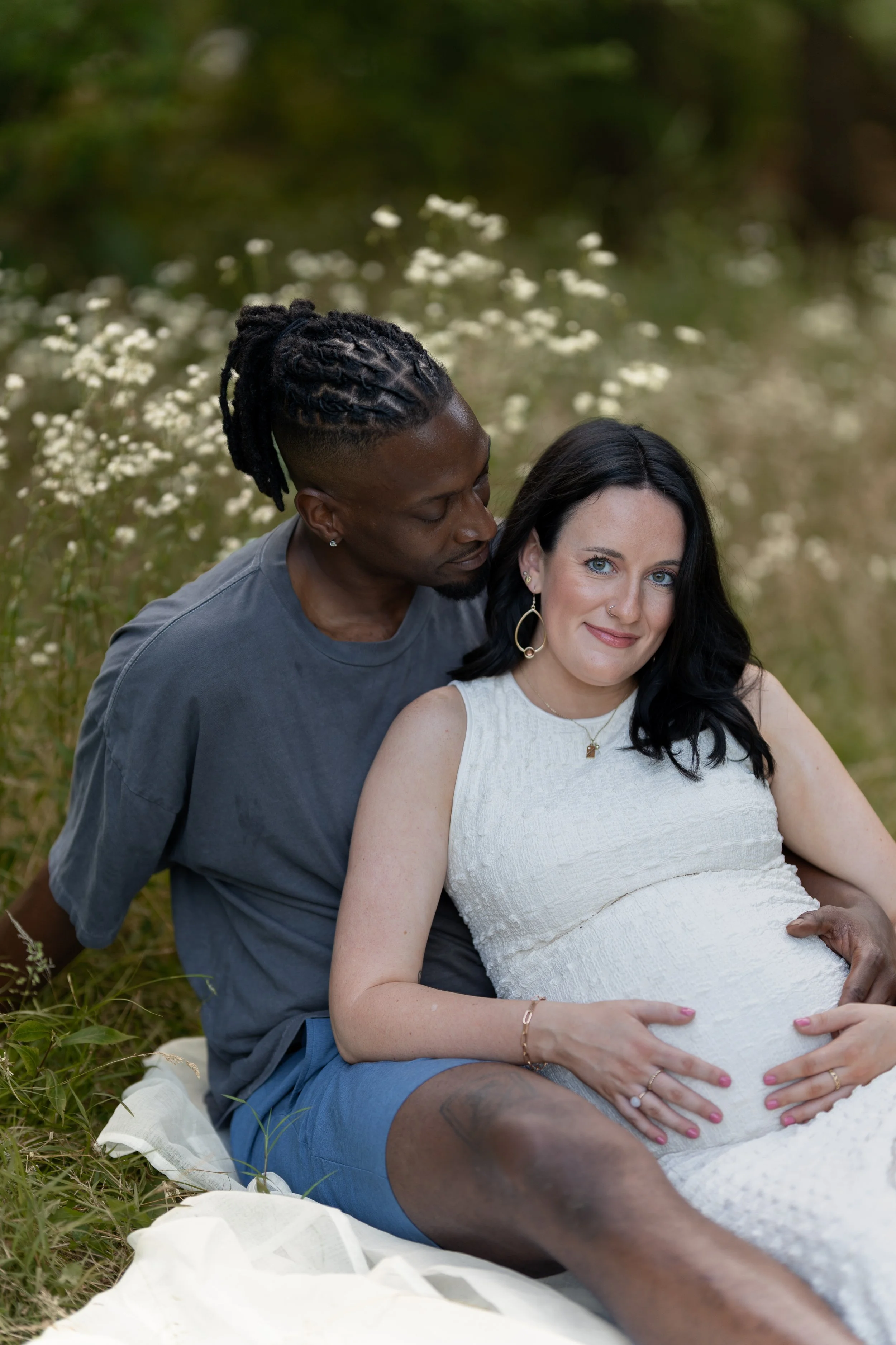 A man with dreadlocks and a woman with black hair, both sitting outdoors in a meadow filled with white flowers. The woman is pregnant, and they are holding her baby bump.