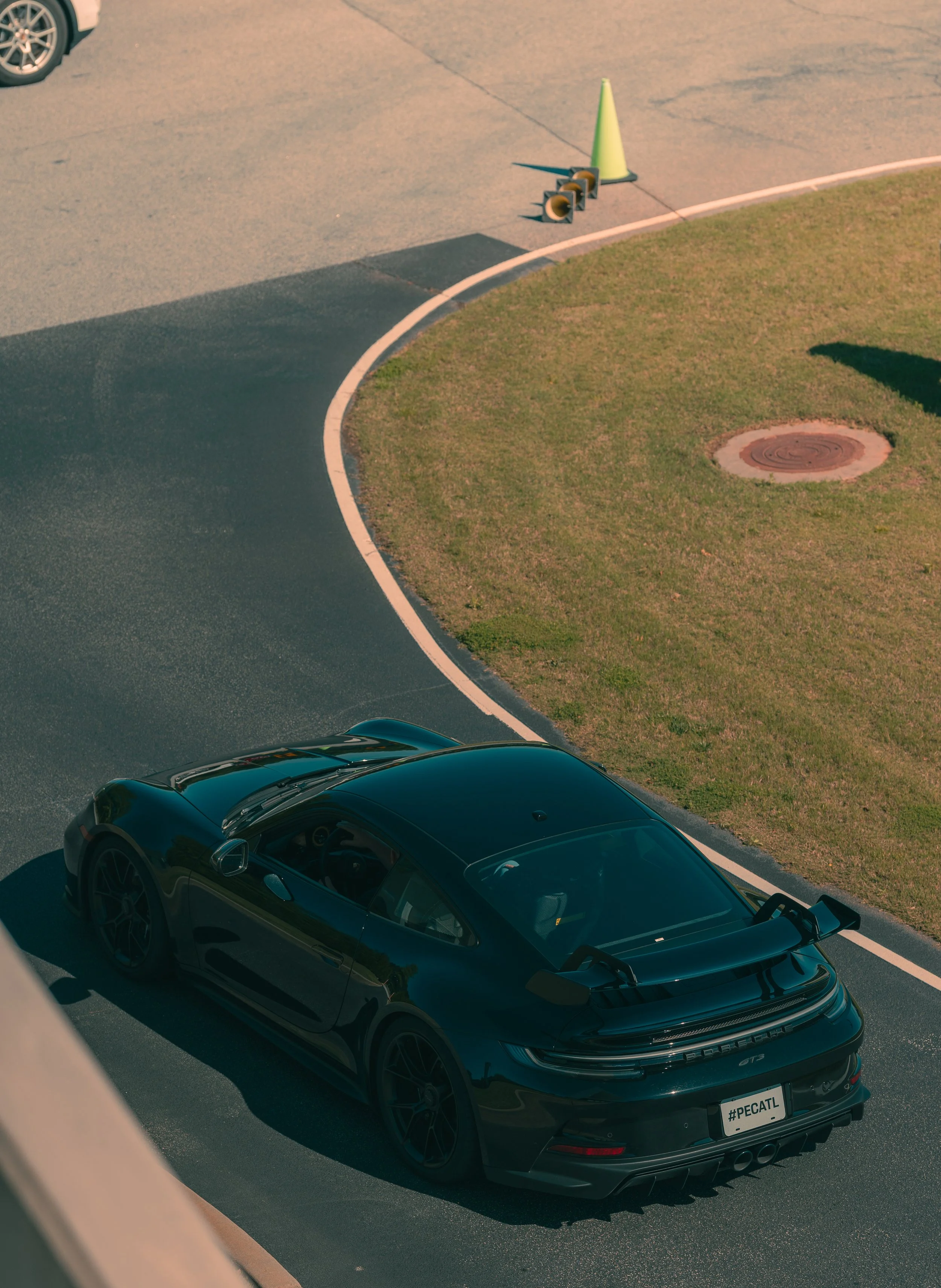 A black Porsche 911 GT3 sports car parked on a paved street next to a grassy area with a manhole cover. A yellow traffic cone and two loudspeakers are on the curb nearby.