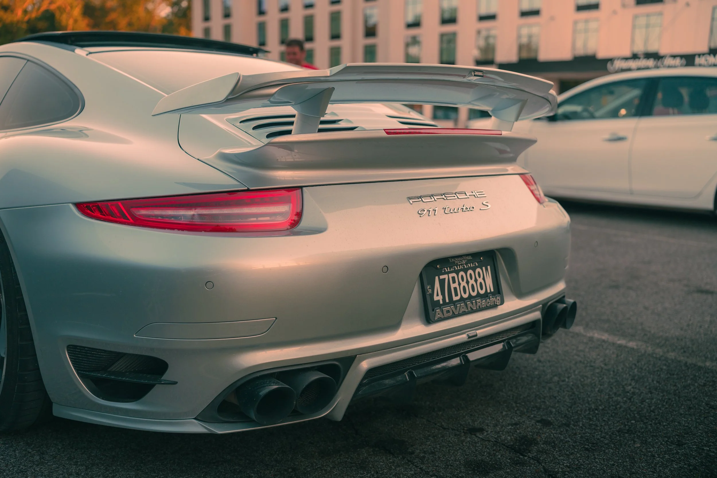 The rear of a silver Porsche 911 Turbo S with a large rear spoiler, dual exhaust pipes, and a California license plate, parked in an outdoor lot with other cars and a building in the background.