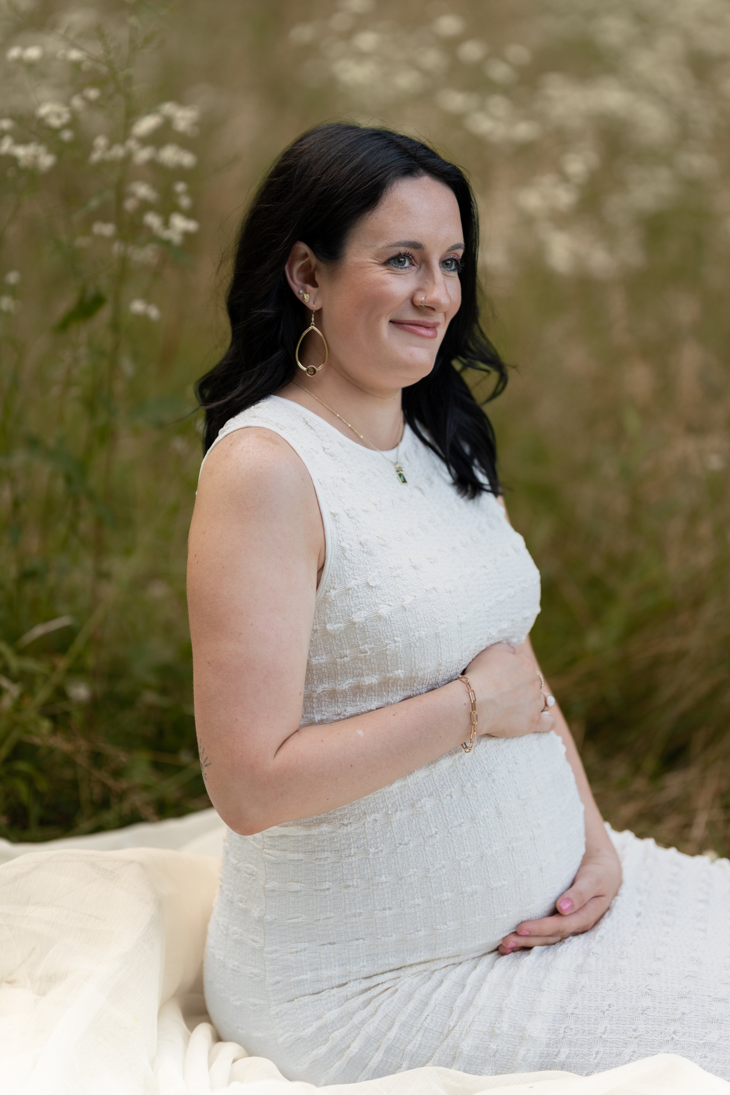 A pregnant woman with dark hair, wearing a white dress, sitting outdoors with her hands on her belly, surrounded by greenery and white flowers.