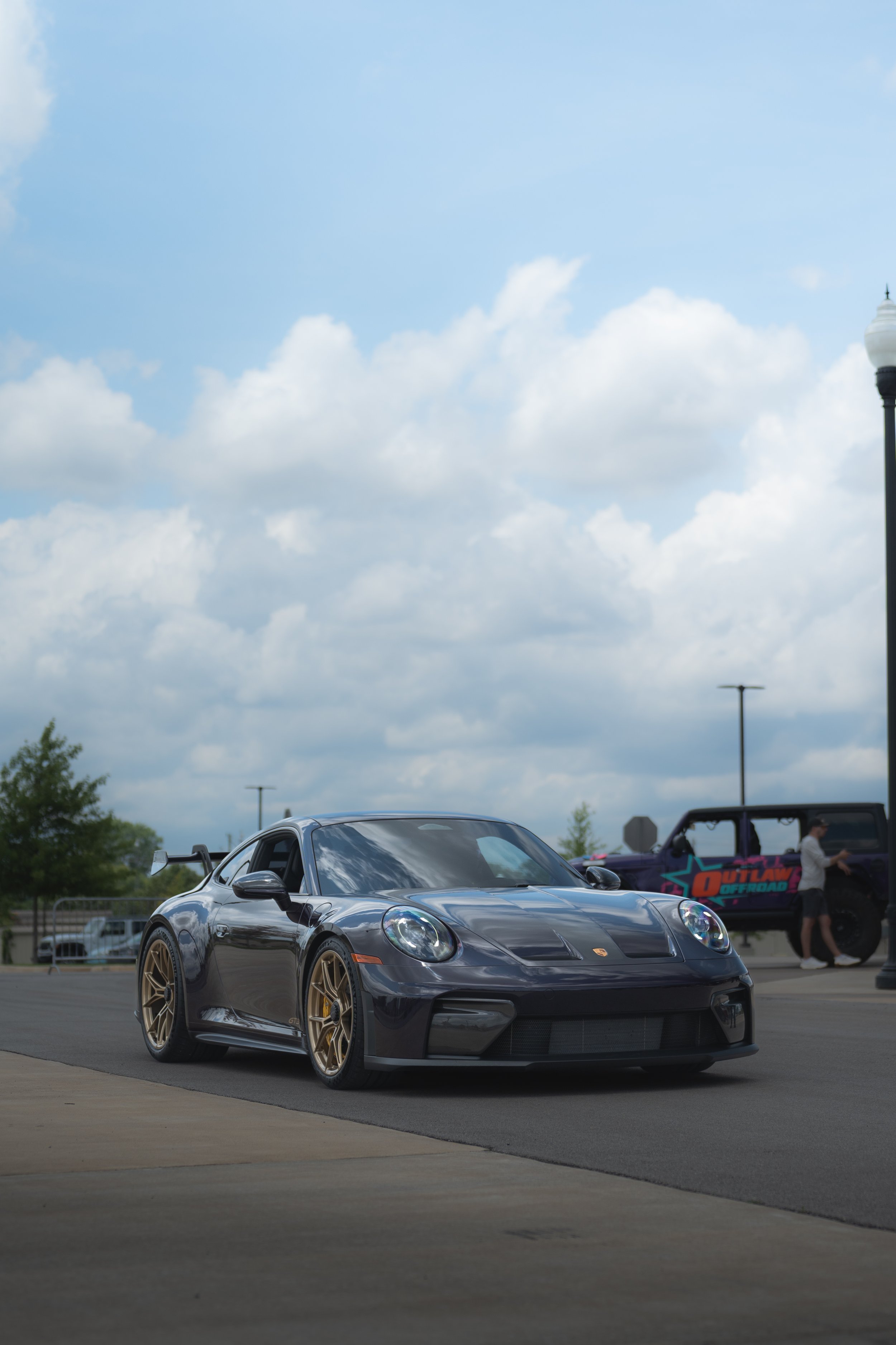 A black sports car with gold rims parked on a paved area outdoors, with a cloudy sky and other vehicles, including a purple off-road vehicle and a person, in the background.