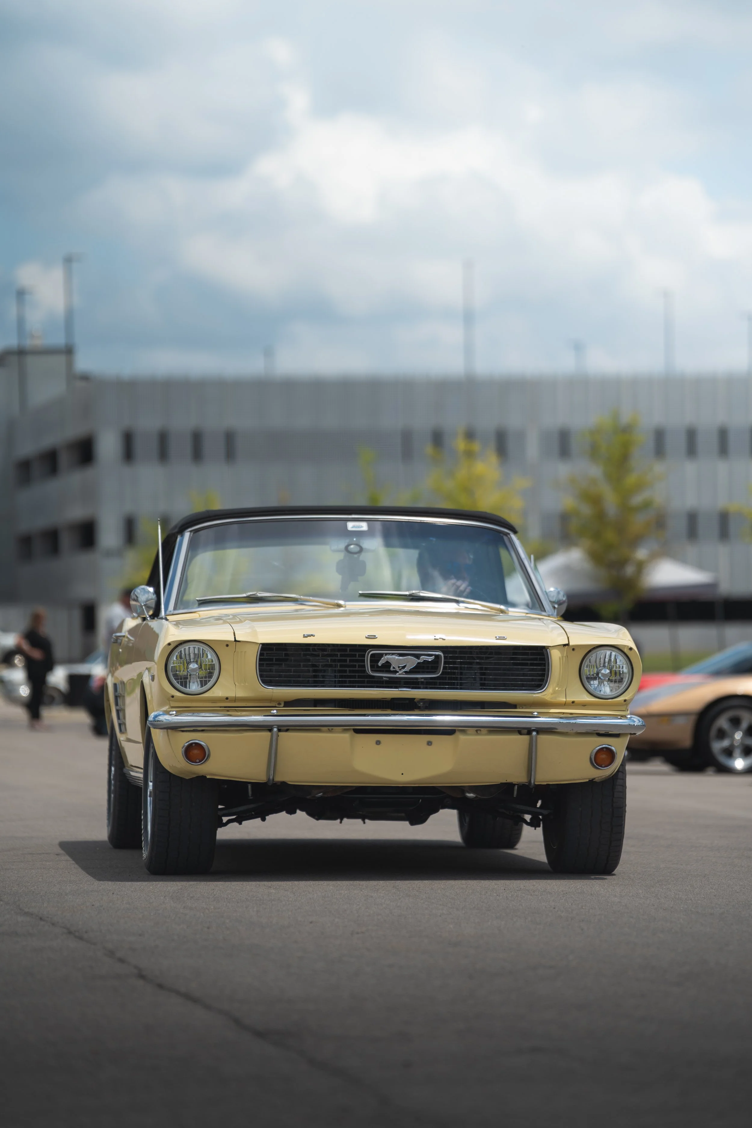 A vintage yellow Ford Mustang car displayed at an outdoor event, with other classic cars and people in the background.