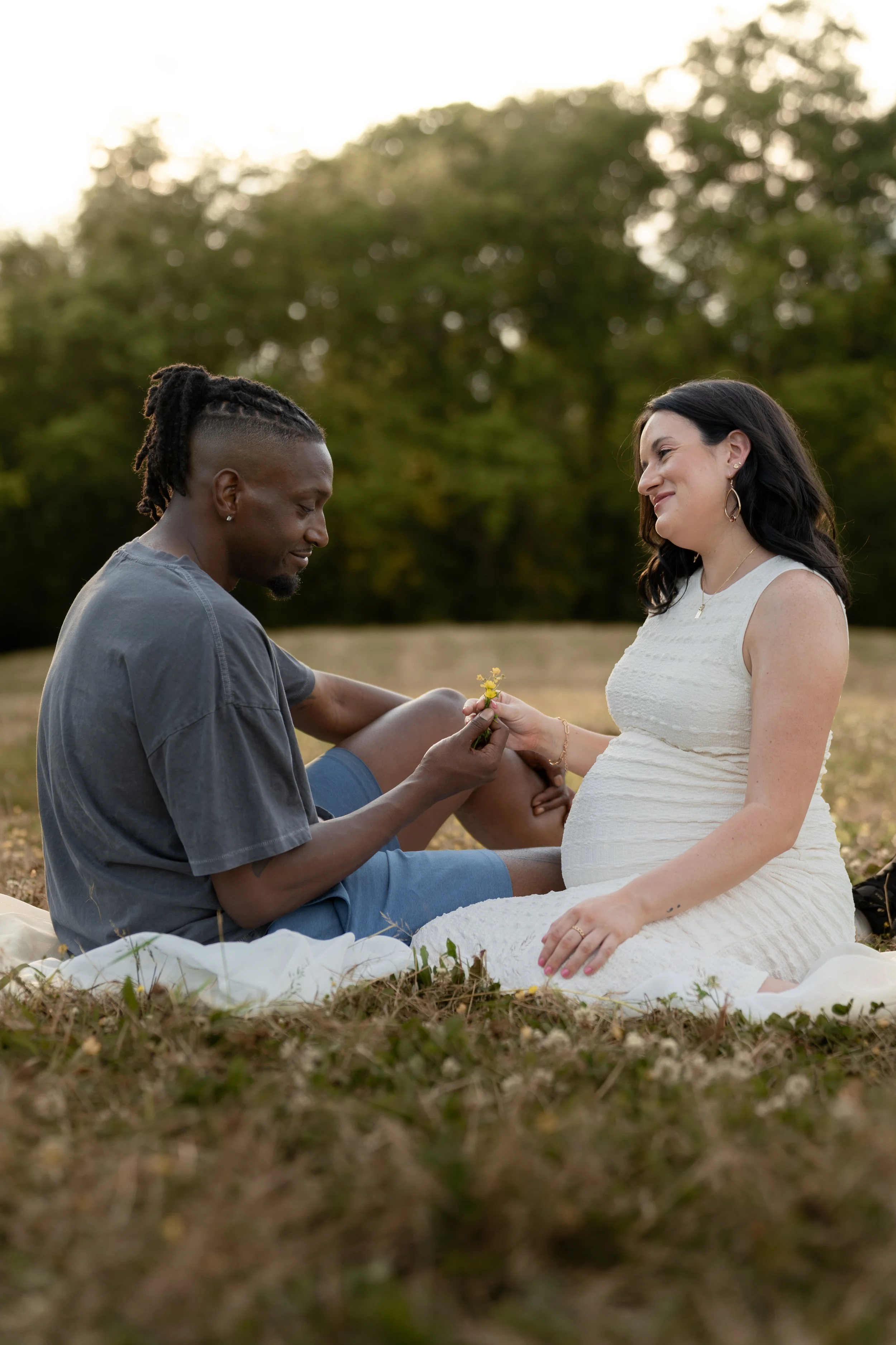 A couple, a man with dark skin and dreadlocks and a woman with light skin and dark hair, sitting outdoors on a blanket, exchanging flowers during sunset. The man is kneeling, and the woman is smiling, wearing a white dress, with a background of green