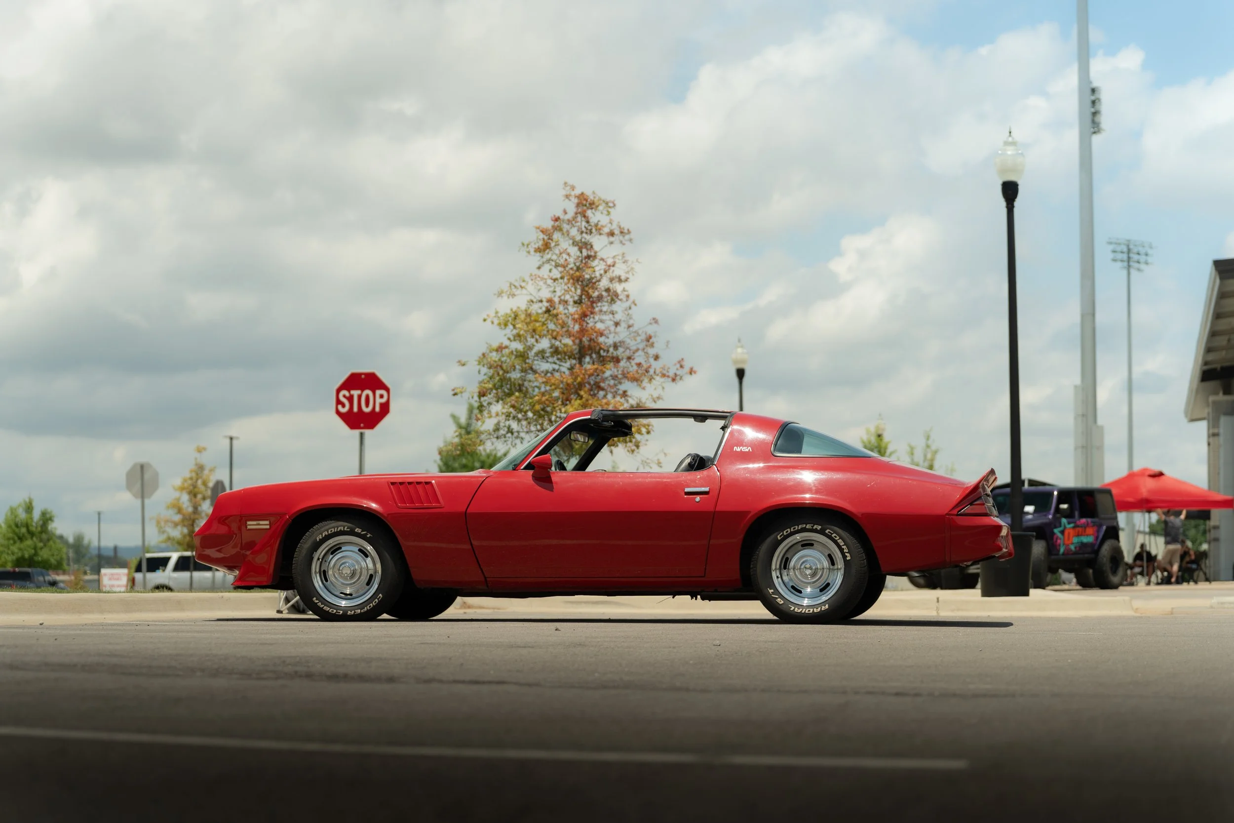 A red vintage sports car is parked in a lot under a cloudy sky, with a stop sign and trees in the background.