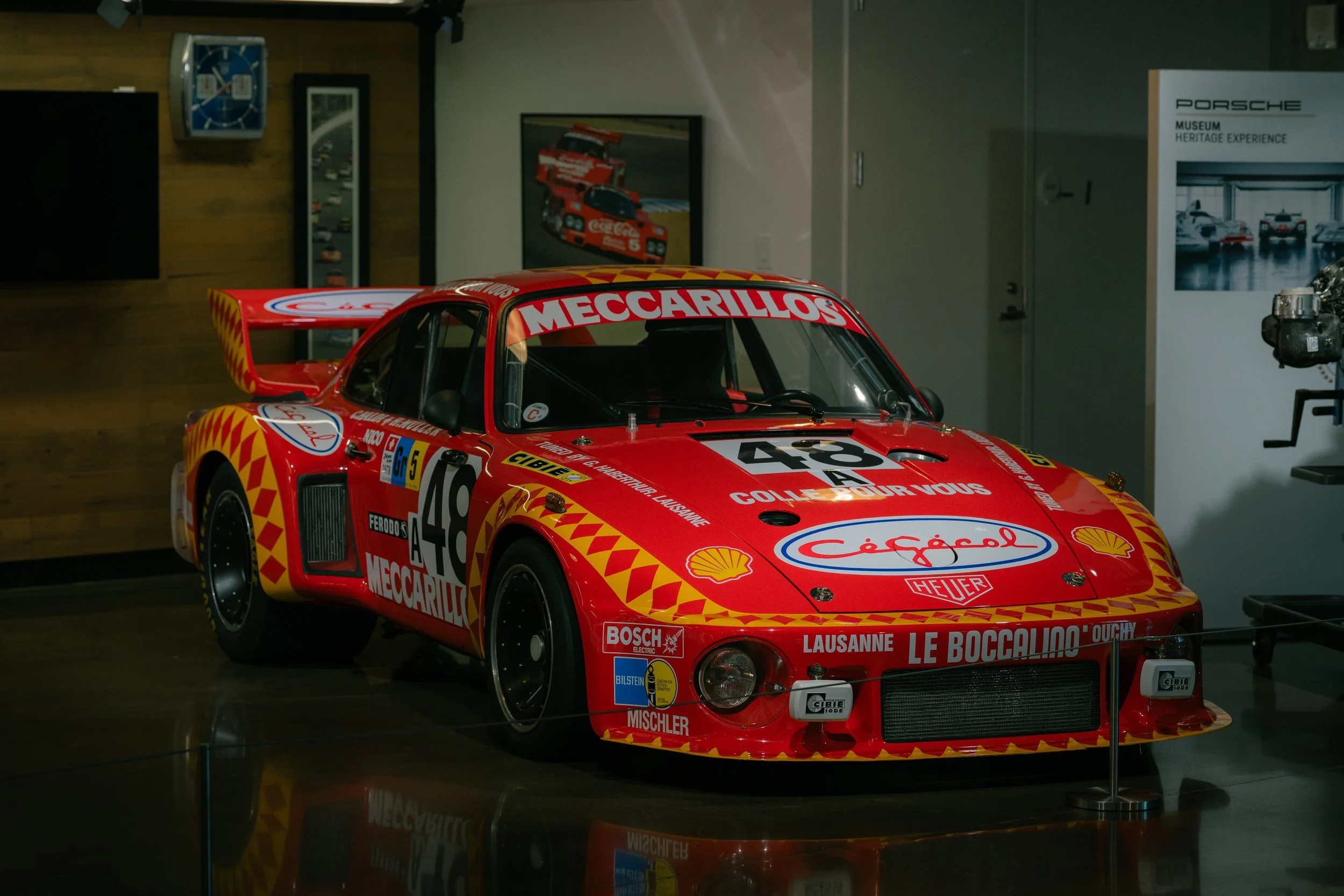 A red vintage racing car displayed in a museum. The car has various sponsor decals and racing number 43, with a large rear wing and open cockpit. There is a framed picture of race cars on the wall behind it.