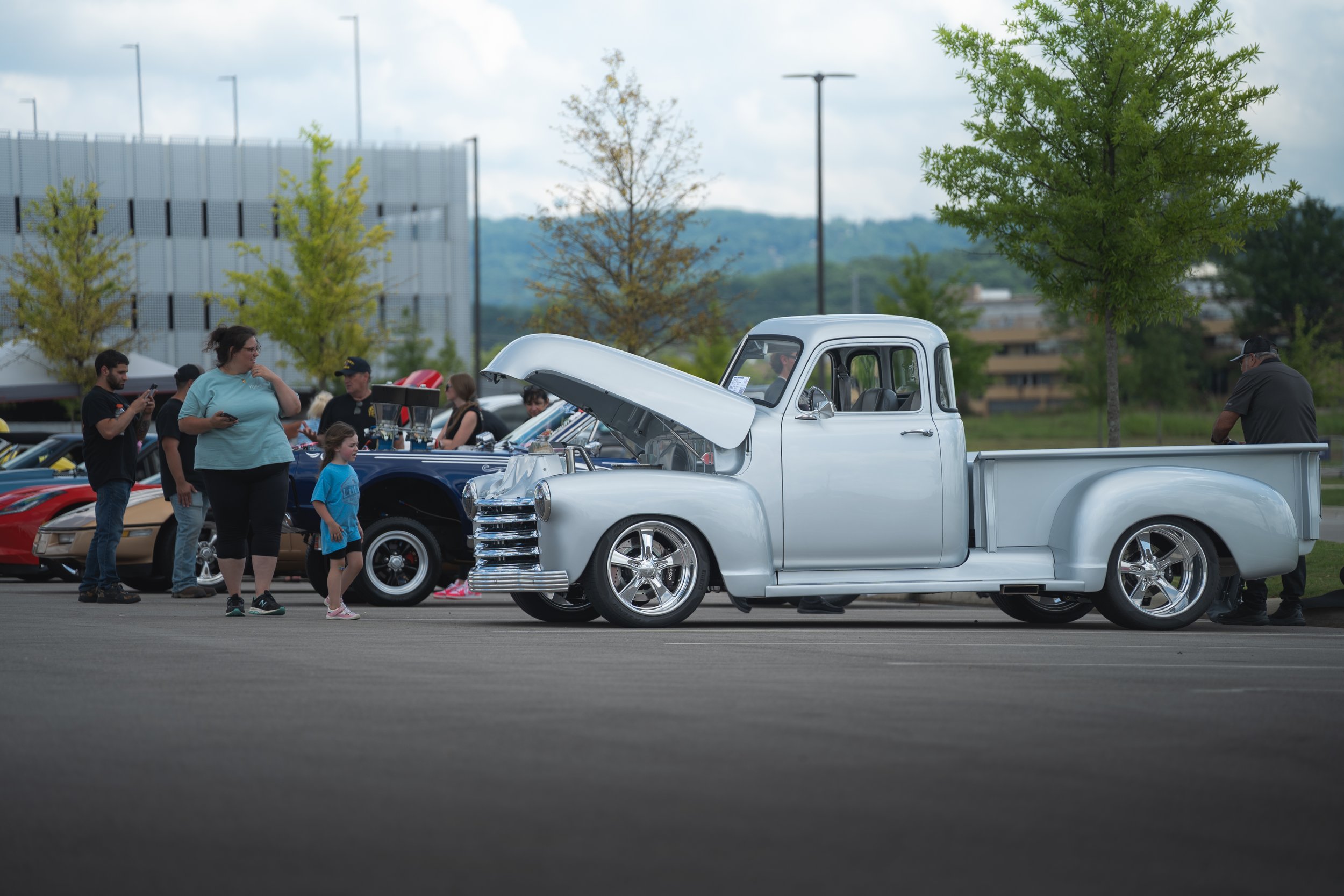 A silver vintage pickup truck at a car show with its hood open, surrounded by people including children and adults, some taking photos. There are other classic cars in the background, trees, and a cloudy sky.